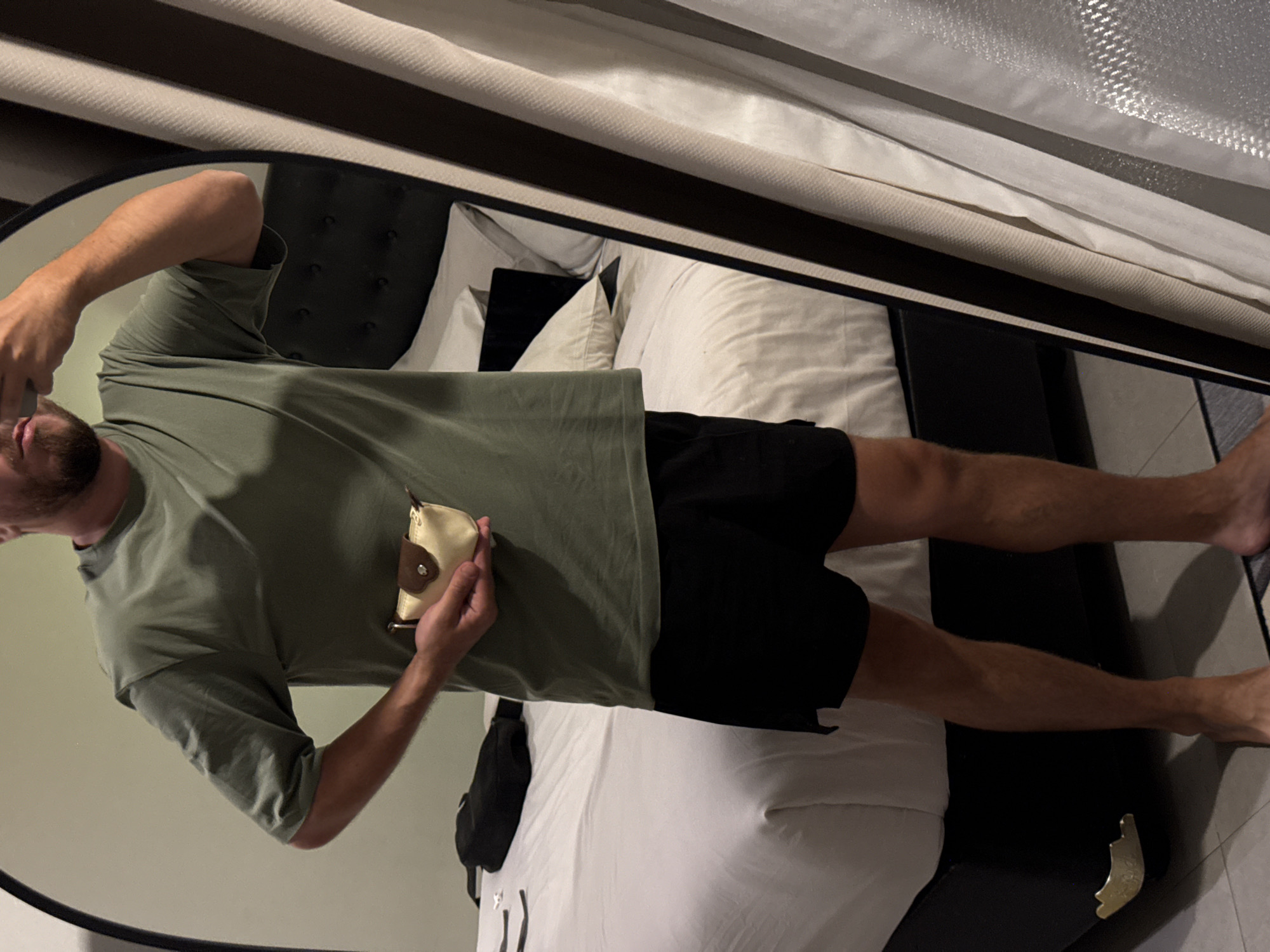 A man in a green t-shirt and black shorts is reflected in a round mirror. He is holding a small, tan-colored pouch. The reflection shows a bed with white sheets and a black headboard. The image appears to be taken indoors, possibly in a hotel room. Based on the request of North Kuta, Indonesia, that cannot be confirmed in the image.