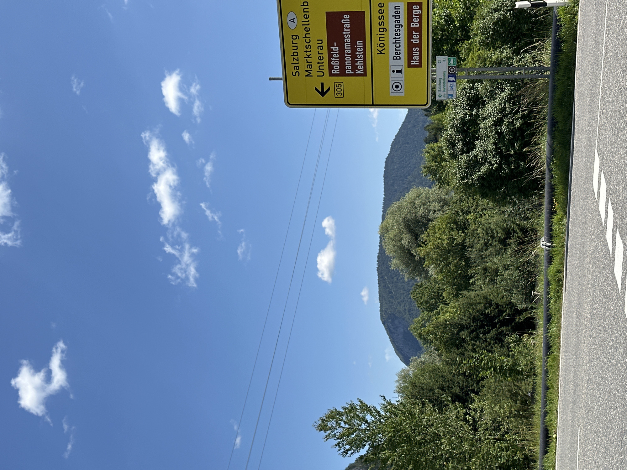 This is a roadside photograph taken on a clear, sunny day. The image features a large yellow directional sign with German text. The sign indicates directions to Salzburg, Marktschellenberg, Unterau, Roßfeld-panoramastraße, Kehlstein, and Königssee. It also points towards Berchtesgaden and "Haus der Berge." Below the main sign, a smaller, older sign displays the number 305 and an arrow pointing left. Further down, another sign indicates the distance to Salzburg (22 km) and Berchtesgaden (8.2 km), along with icons for a visitor center and a post office.
In the background, a lush green forest covers a gentle mountain slope under a bright blue sky dotted with white, fluffy clouds. Power lines are visible stretching across the upper portion of the frame. The foreground shows a portion of a paved road with white lane markings. A black pipe runs along the edge of the road, bordered by green grass and vegetation.