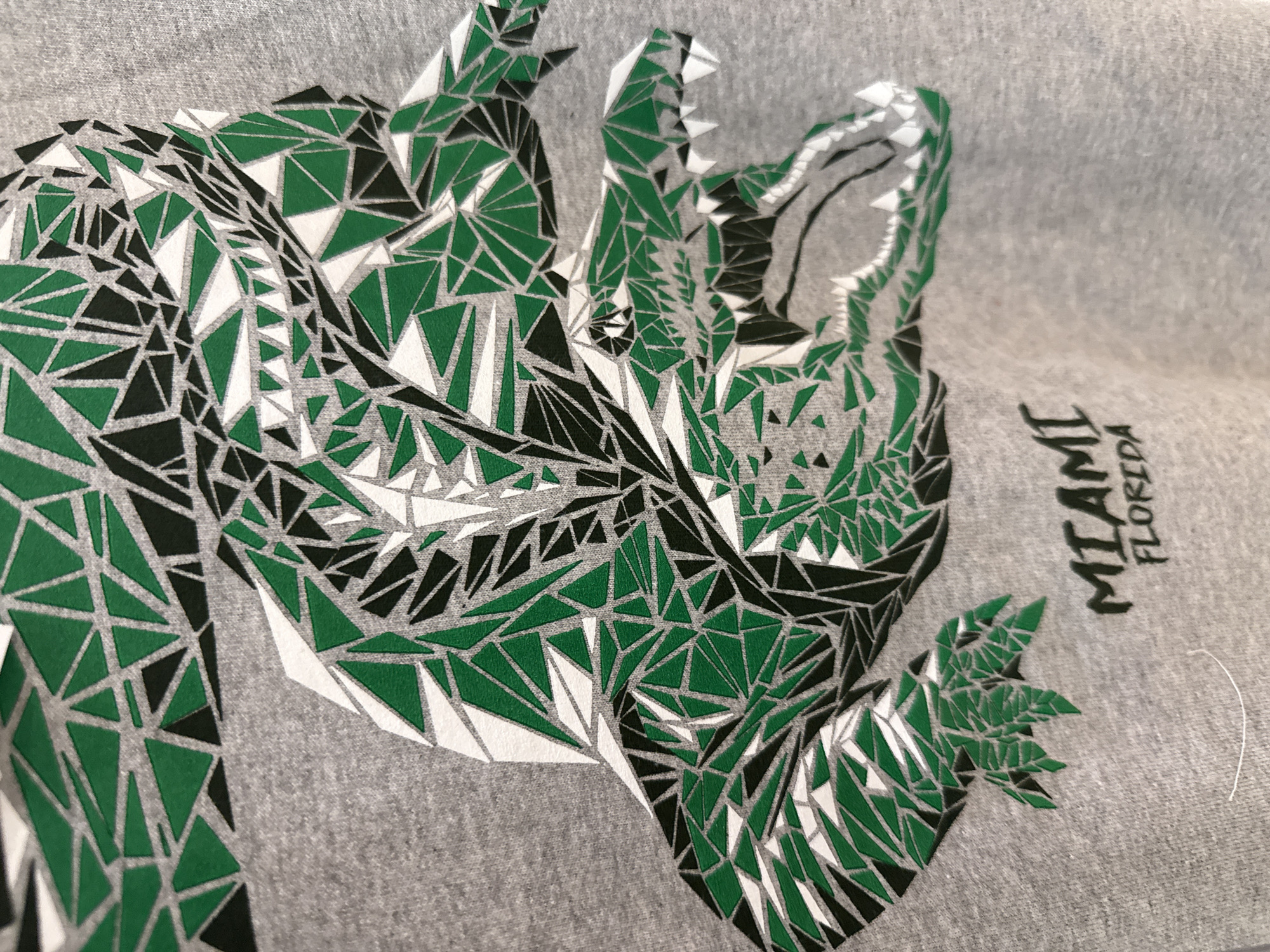 The image shows a gray T-shirt with a geometric design of a reptile, possibly an alligator or crocodile. The design is composed of small triangles in green, black, and white. To the right of the reptile design, the words "MIAMI FLORIDA" are printed vertically. The location of the scene is not specified.