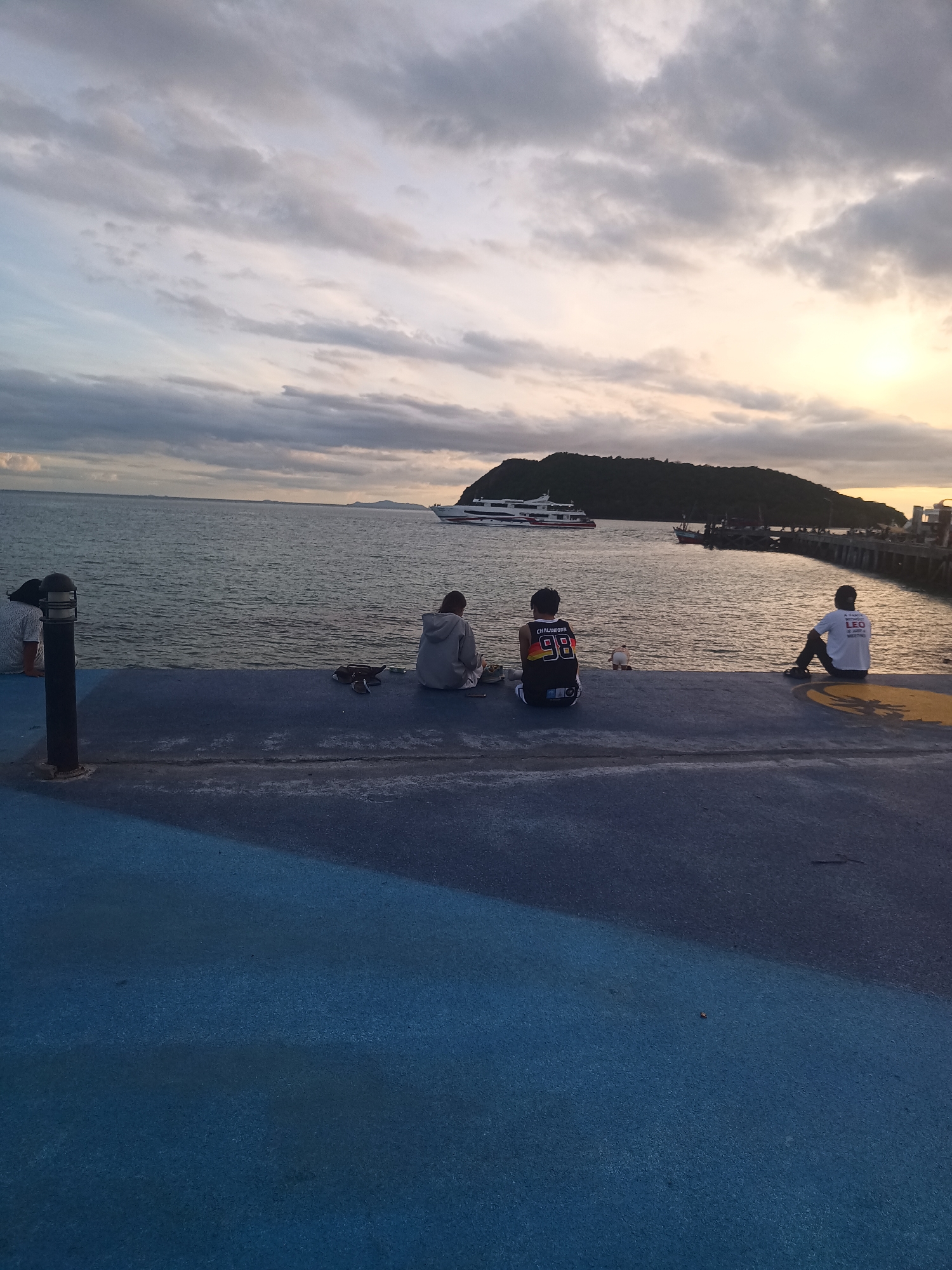 The photo captures a scene at dusk in ตำบลเกาะพะงัน, Thailand. Several people are sitting along a concrete pier that extends into the water. From left to right, there is a person sitting out of view near a lamp post; a person in a grey hoodie sitting on the pier; a person wearing a Chaladonn 98 basketball jersey also sitting on the pier; and a person in a white t-shirt with the word "LEO" sitting at the end of the pier. In the background, there is a body of water with a large white ferry or boat and a smaller fishing boat. A green, hilly island is visible in the distance. The sky is filled with clouds tinged with orange from the setting sun. The pier has a painted blue surface.