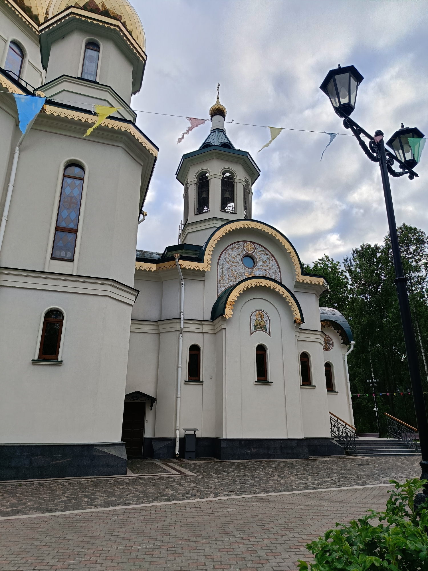 The image depicts the Church of the Holy Great Martyr Catherine in Murino, Saint Petersburg, Russia. The church is constructed in a traditional Orthodox style with white walls, brown trim, and green and gold domes. Pennant flags decorate the space. A black street lamp is present to the right of the image. The sky is cloudy. The foreground shows a brick paved plaza.