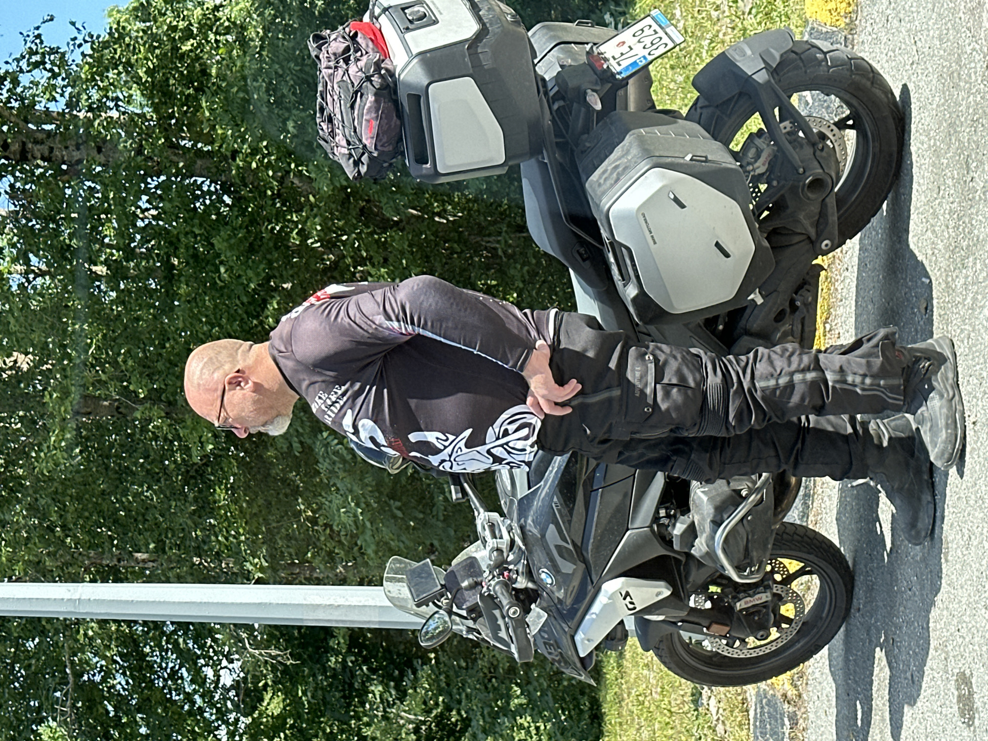 A middle-aged, bald, Caucasian male with glasses and a goatee is standing next to a black BMW GS motorcycle. He is wearing a black long-sleeved shirt with a white graphic design and black riding pants with white accents. The motorcycle is equipped with saddlebags and a top case, and a red and black bag is strapped to the top of the top case. The motorcycle has a license plate that reads "7E 3629". The man is looking to his right, with his hands on his hips. The motorcycle is parked on a paved surface next to a grassy area. In the background, there are trees and a light pole. The scene appears to be outdoors on a sunny day. The location is identified as Crkvine, Montenegro.