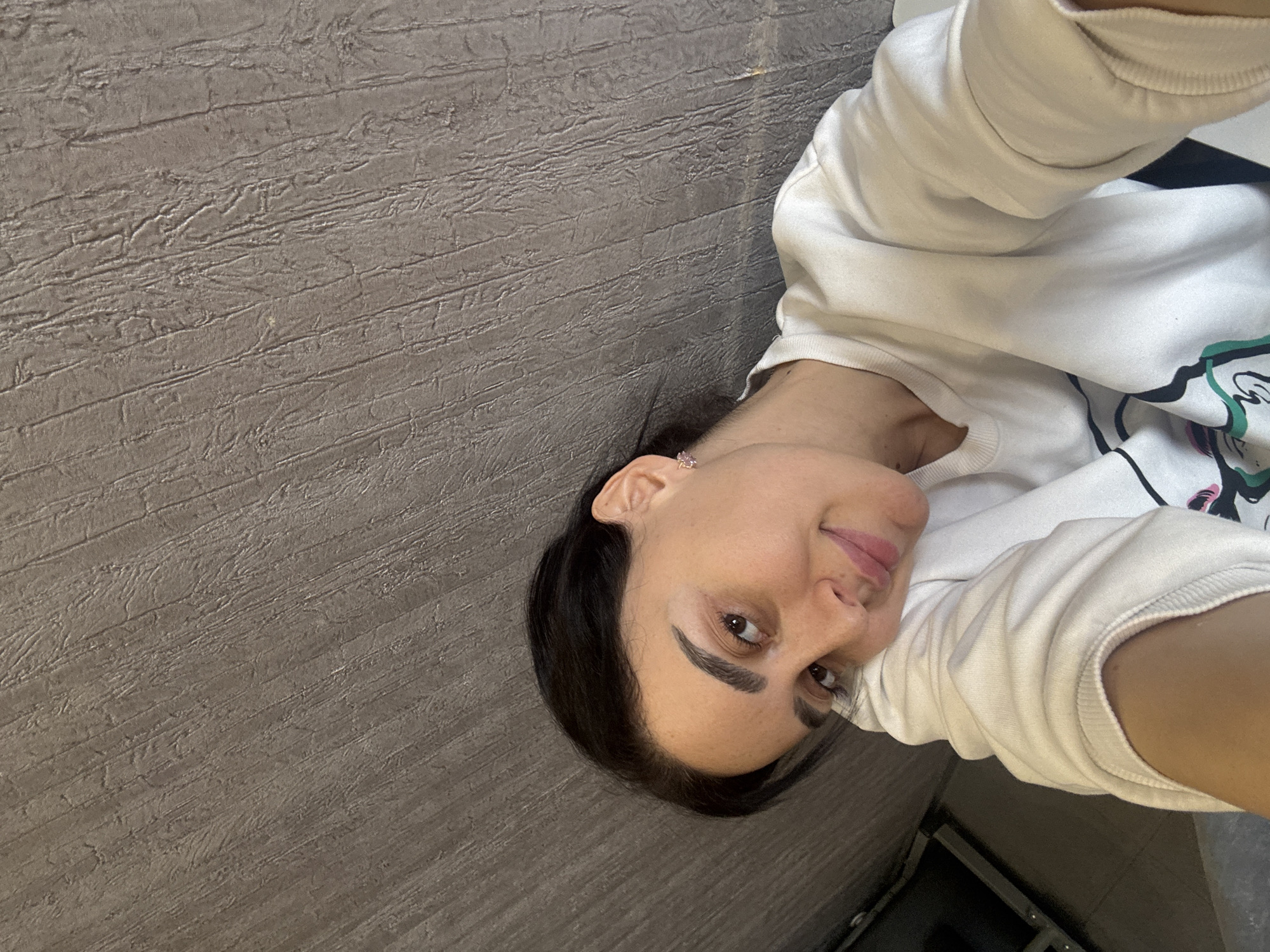 A woman is captured in a close-up photograph, likely a selfie, set against a textured grayish-brown wall in an indoor environment, with the context of Tbilisi, Georgia. The image is oriented 90 degrees counter-clockwise.
The subject is a woman with dark hair, prominent dark eyebrows, and brown eyes, looking directly towards the camera with a gentle, slightly smiling expression. She is wearing a white, long-sleeved sweatshirt, on which a multi-colored graphic design is partially visible. A small, light-colored earring can be seen in her left ear.
The background features a distinctive wall with horizontal lines and a rough, tactile texture, reminiscent of grooved wood or a stucco finish. The lighting suggests an indoor setting. No other individuals or objects are clearly visible, and there is no legible text within the frame. The overall atmosphere conveyed is casual and relaxed.