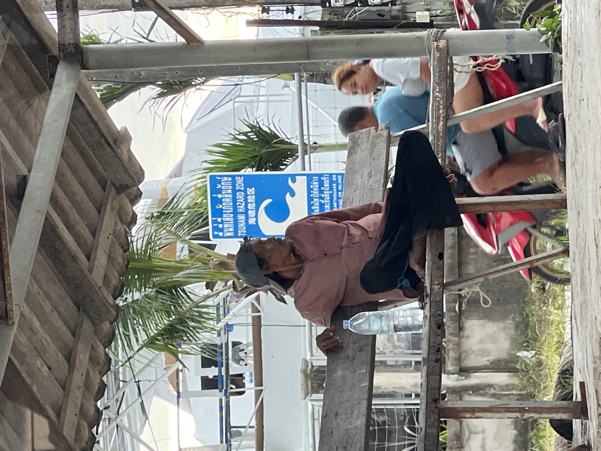 In Chalong, Thailand, a man wearing a pink shirt and a grey baseball cap sits on a weathered wooden bench under a rustic roof structure. A plastic bottle of water is placed beside him on the bench. To his right, a blue sign indicates a "Tsunami Hazard Zone" with wave graphics and text in Thai and Chinese. In the background, palm trees are visible, along with parts of buildings and a large white tent-like structure. Further to the right, two other individuals are partially visible, one wearing a blue shirt and the other a white top, seated on another bench or a motorcycle. A red motorcycle is parked in front of them. The ground appears to be paved, with overgrown grass and some debris in the foreground. The overall scene suggests an outdoor public area, possibly a waiting area or a rest stop.