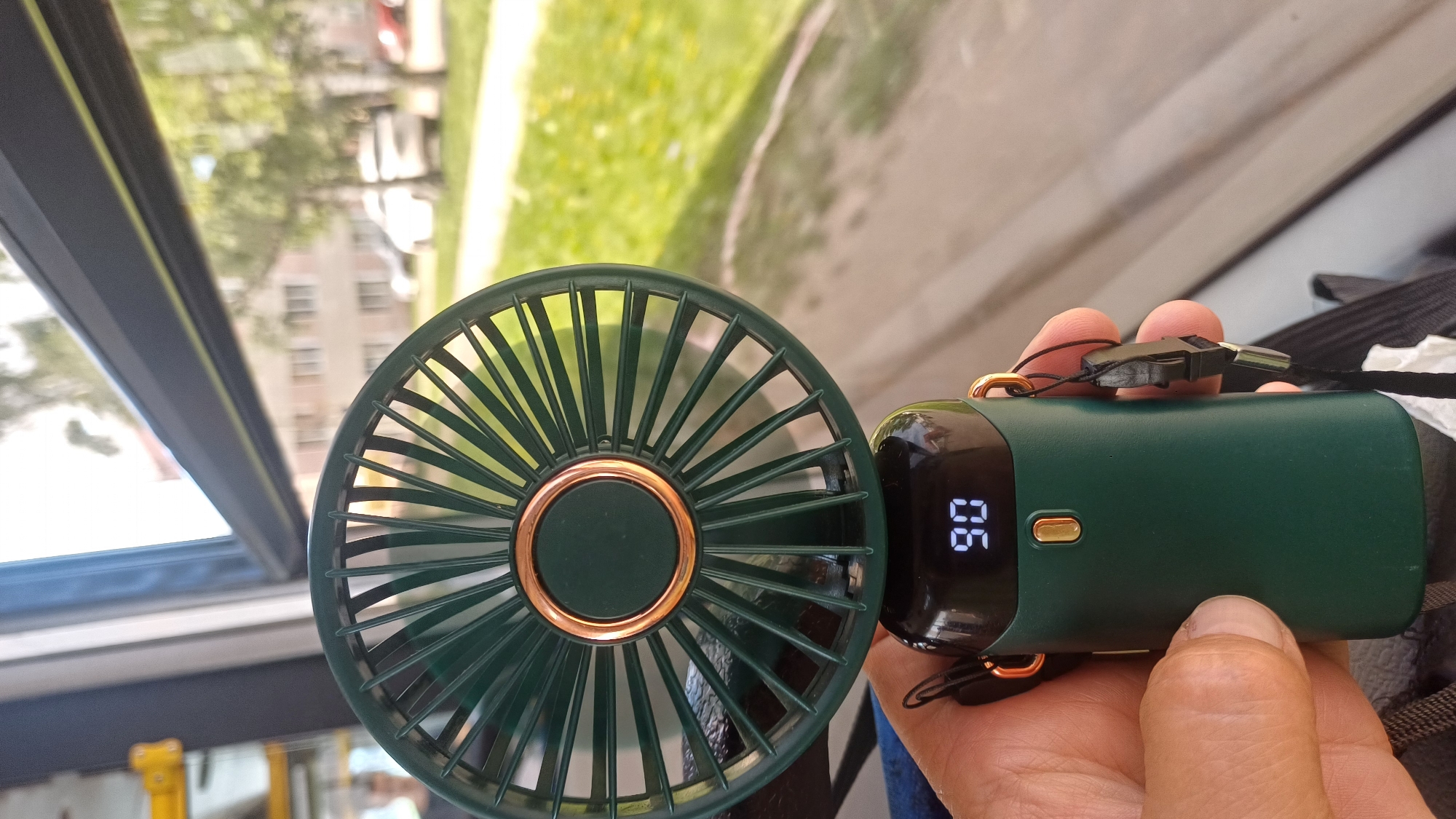A close-up shot captures a person's hand holding a portable, dark green electric fan. The fan has a metallic gold ring around its center and a digital display showing "90" in white illuminated digits. The fan is positioned in front of a window, through which a blurred view of an urban environment is visible. The background suggests a daytime scene with buildings and trees outside, likely in Izhevsk, Russia, given the context. The overall lighting is natural, indicating the photo was taken during daylight hours.