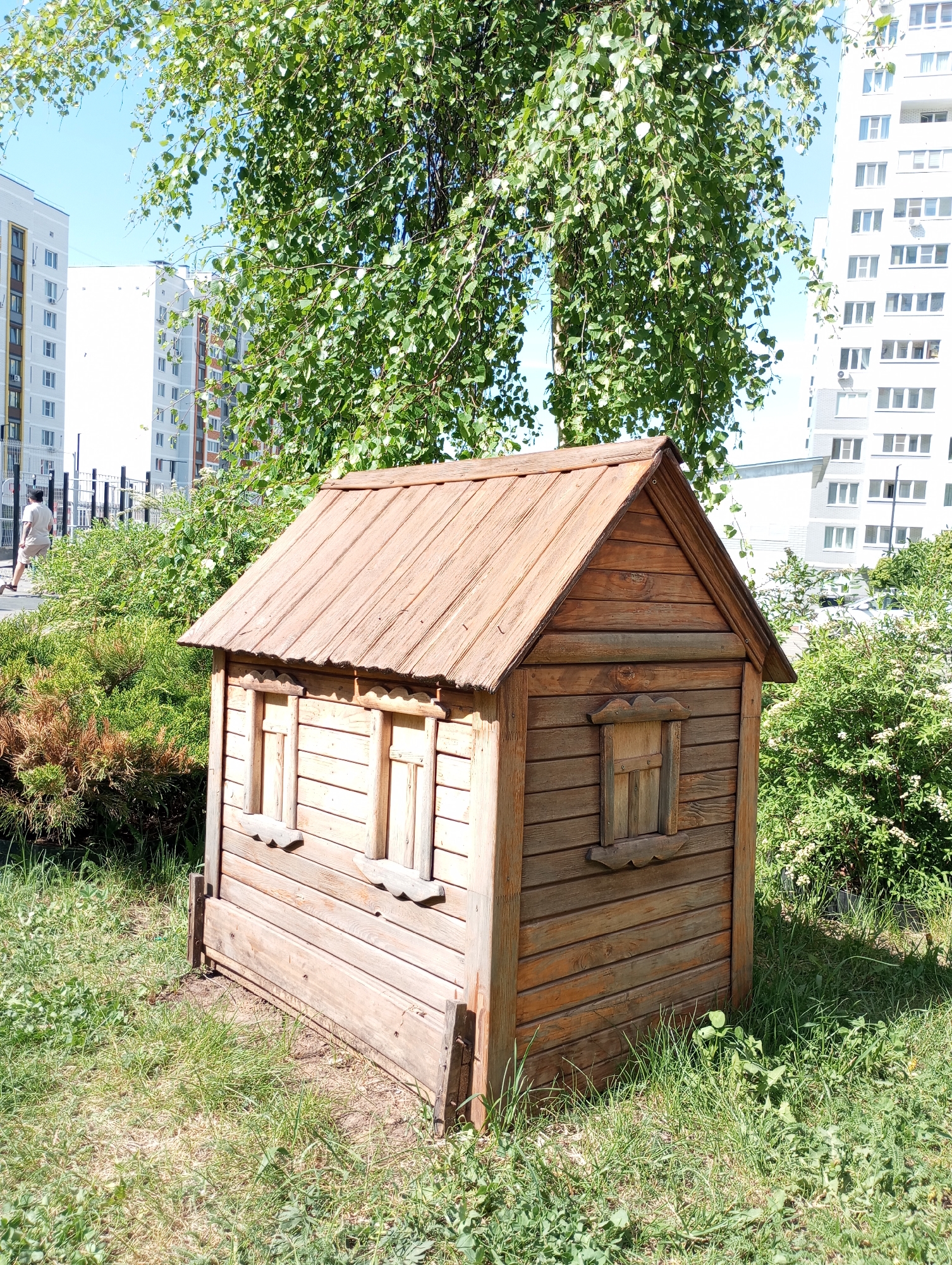 The image depicts a small, wooden playhouse situated in a grassy area. The playhouse has a gabled roof, and windows with decorative trim. Behind the playhouse, there are bushes and trees, with tall white apartment buildings visible in the background. A person can be seen walking in the distance on a path near a fence. The image was taken in Izhevsk, Russia.