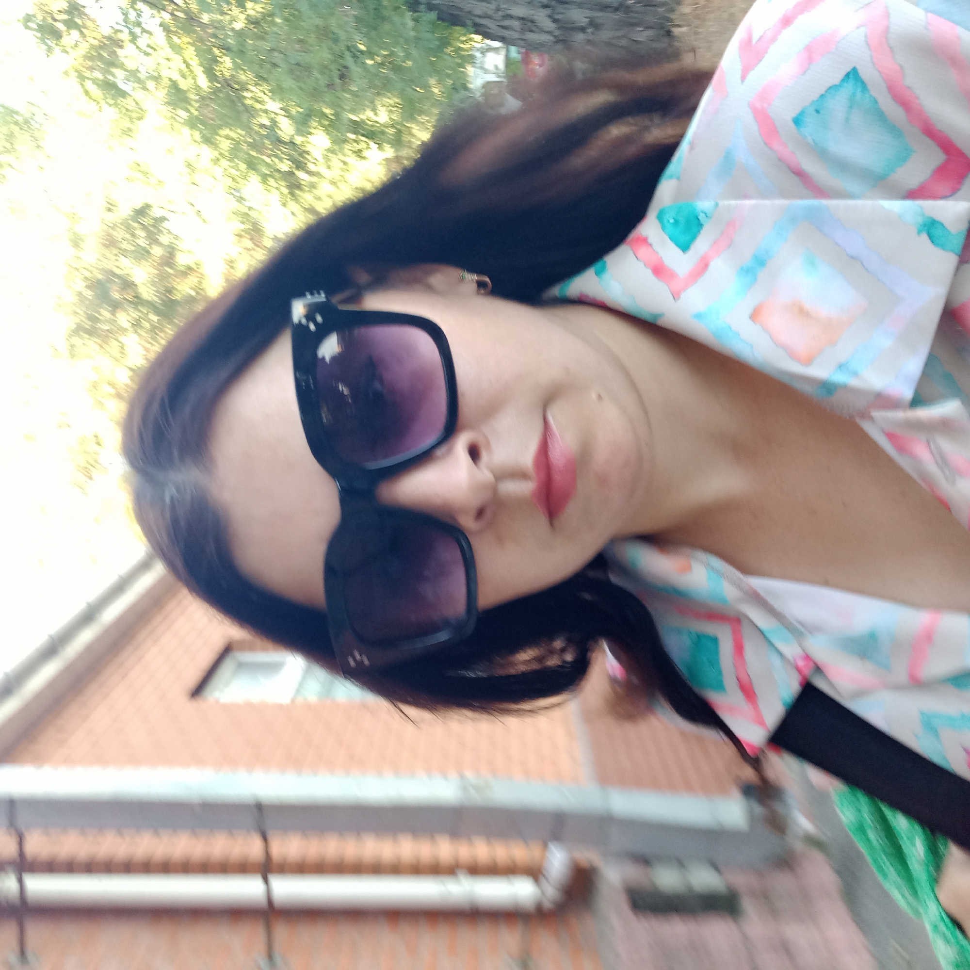 A medium shot image shows a woman wearing oversized dark sunglasses and a light-colored jacket with a diamond pattern. She has dark hair and is looking at the camera. In the background, a brick building with a downspout is visible on the left, and trees with green foliage are on the right. The image appears to be taken outdoors during the day. The exact location in Kropyvnytskyi, Ukraine cannot be determined from the visual information alone.