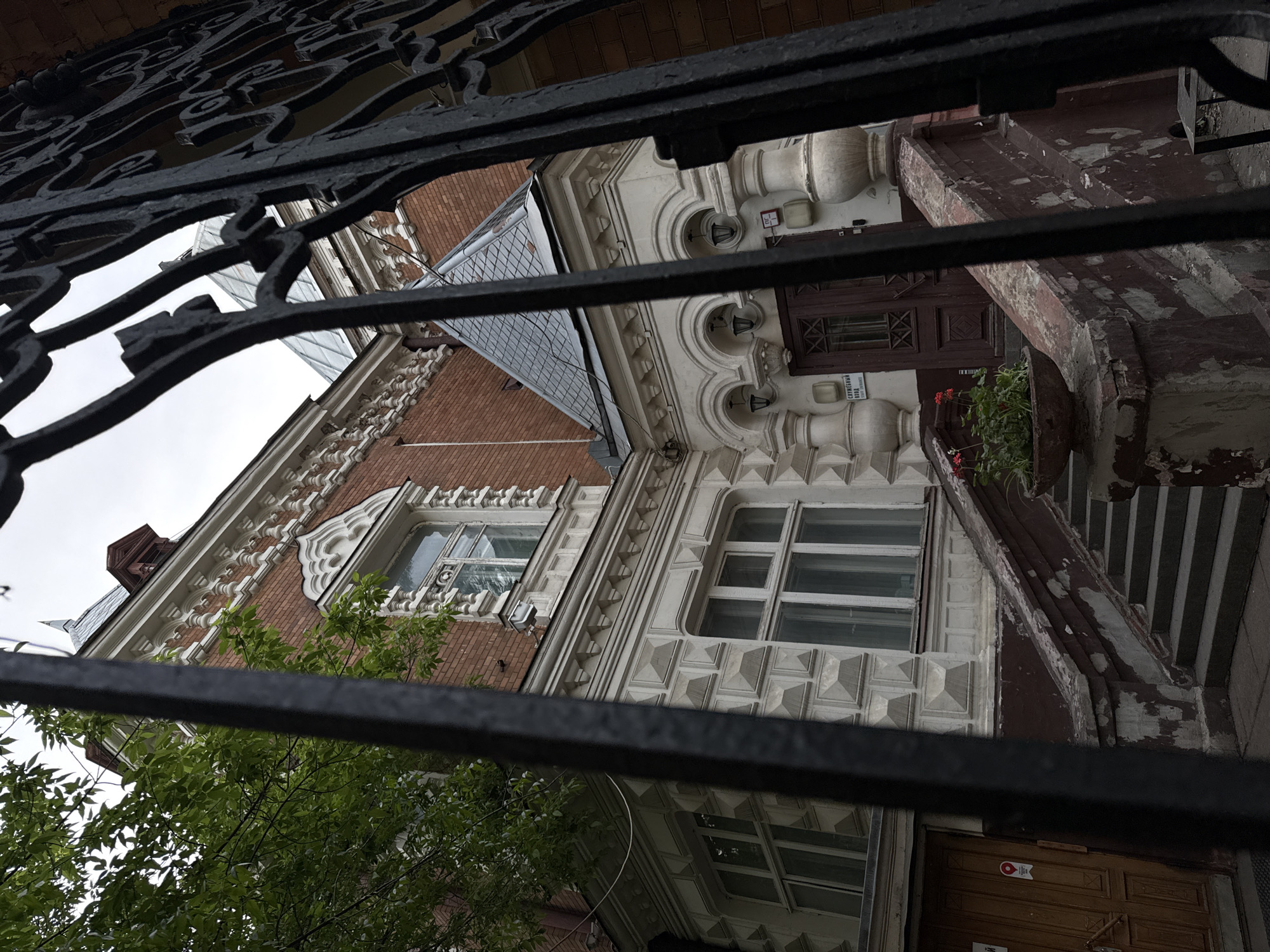 An eye-level shot of an ornate building facade viewed through a wrought-iron fence. The facade features a mix of red brick and white architectural elements, with decorative details around the windows and roofline. A grey slate roof section is visible. Some green foliage from a tree or shrub is visible on the left side of the image. The fence is black, and its intricate design partially obscures the view of the building. The building features a raised entry way with a brown wooden door. The picture was taken in Moscow, Russia.
