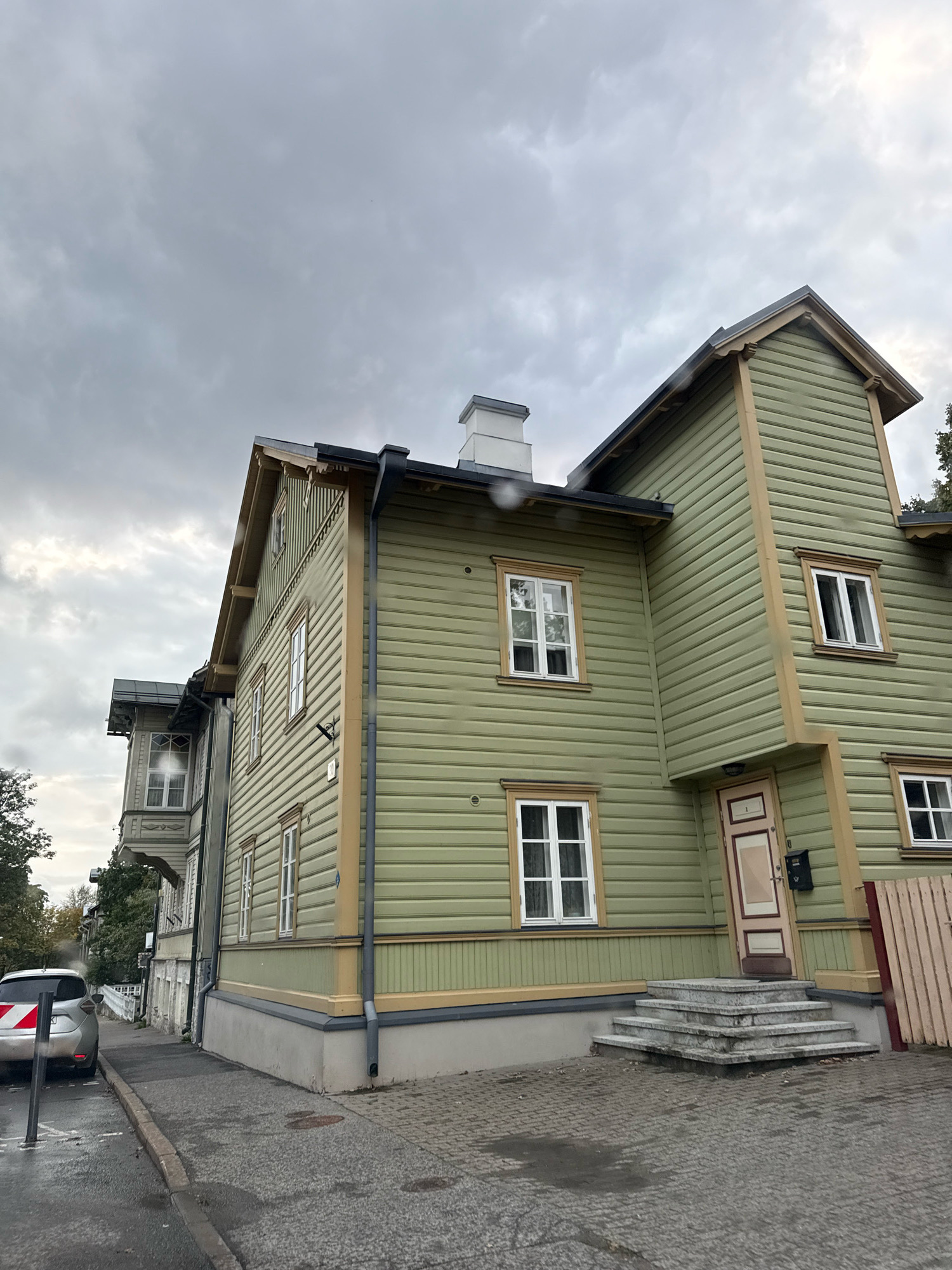 This is a photograph of two adjoining wooden houses in Tallinn, Estonia. The house in the foreground is painted light green with yellow trim and features a prominent staircase leading to the entrance. The house to the left is a lighter color, possibly white or cream, and has more ornate architectural details. A silver car with a reflective red and white striped marker is parked on the wet street to the left of the frame. The sky is overcast and gray, suggesting a cloudy or rainy day. The street in front of the houses is paved with cobblestones and asphalt. The overall scene depicts a residential street in an urban environment with historic architecture.