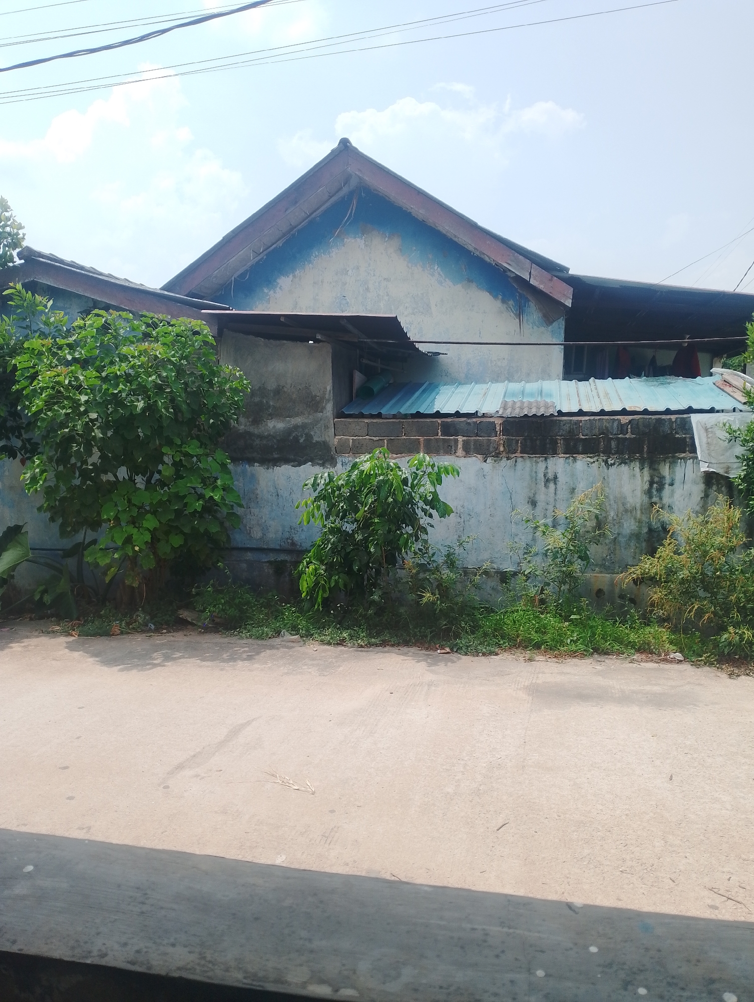 The image shows a street-level view of a building and its surroundings in Batam, Indonesia. The building appears to be a single-story structure with a light blue and white facade, a weathered appearance, and a corrugated metal roof. A low wall, constructed of concrete blocks, separates the building from the street. Vegetation, including trees and shrubs, grows along the wall and in front of the building. A paved road is visible in the foreground. The sky is visible in the upper portion of the image, with some clouds present. There are no people or vehicles depicted in the image. The overall impression is one of a residential or commercial area in a tropical climate.