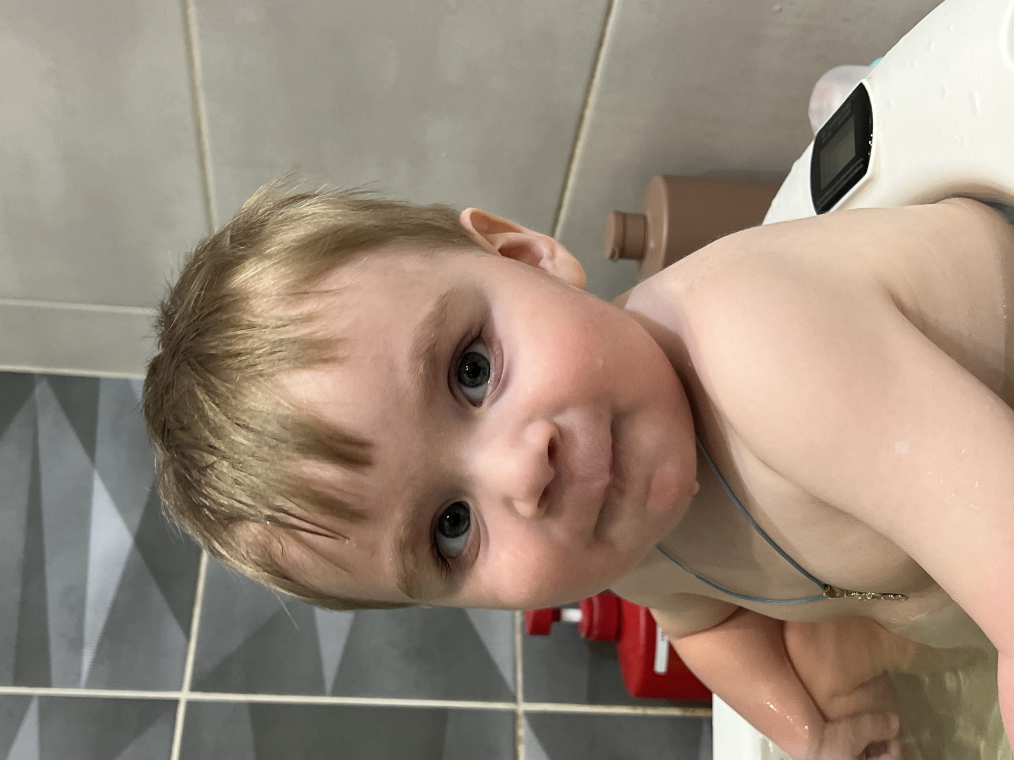 A young child, likely a toddler, is captured in a close-up portrait while taking a bath. The child has light blonde or light brown hair, which appears wet, and striking blue-green eyes. Their expression is neutral to slightly curious, looking directly at the viewer. A thin, light blue cord necklace with a small, indistinct pendant is visible around their neck.
The child is partially submerged in water within a white bathtub or baby bath seat, a portion of which is visible on the right side of the frame. A small black digital display is present on the white bath seat, but any specific text or numbers are unreadable. In the background, a light brown/tan pump bottle and a red container are visible. The bathroom walls are tiled, with a pattern of grey geometric shapes on the lower left and plain lighter grey tiles on the upper sections. The image is taken from a slightly elevated, tilted perspective. The scene depicts a bath time moment in Noviy, Russia.