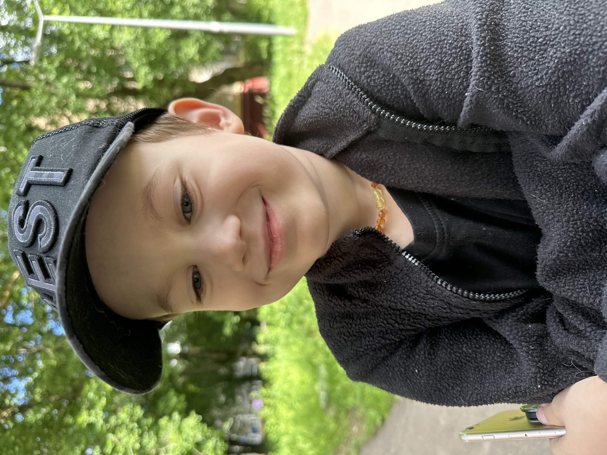 A young boy, likely of Caucasian descent and appearing to be under ten years old, is captured in a close-up, vertically oriented outdoor shot. He is wearing a black baseball cap with prominent grey lettering, partially obscuring his forehead, and a fuzzy black zip-up jacket over a black t-shirt. A multi-colored beaded necklace, with amber and lighter colored beads, rests on his collarbone. His face is turned towards the camera, and he has a gentle smile, with his eyes looking slightly to the side. The background is blurred but suggests a green, leafy environment, likely trees and foliage, indicating an outdoor setting during the daytime. The lighting suggests it is a sunny day. The boy's expression is calm and happy.
The camera appears to be held by someone else, as a hand holding a silver smartphone is visible in the lower right corner of the frame. The phone has a black circular object attached to its back, possibly a PopSocket or a similar accessory. The scene is likely taking place in Moscow, Russia, given the provided location context. No specific event is discernible, but the overall impression is one of casual leisure in a park or garden. The image's orientation is rotated, with the boy's head appearing at the top of the frame when viewed normally. The focus is on the boy's face and upper body, with the background serving to provide context.