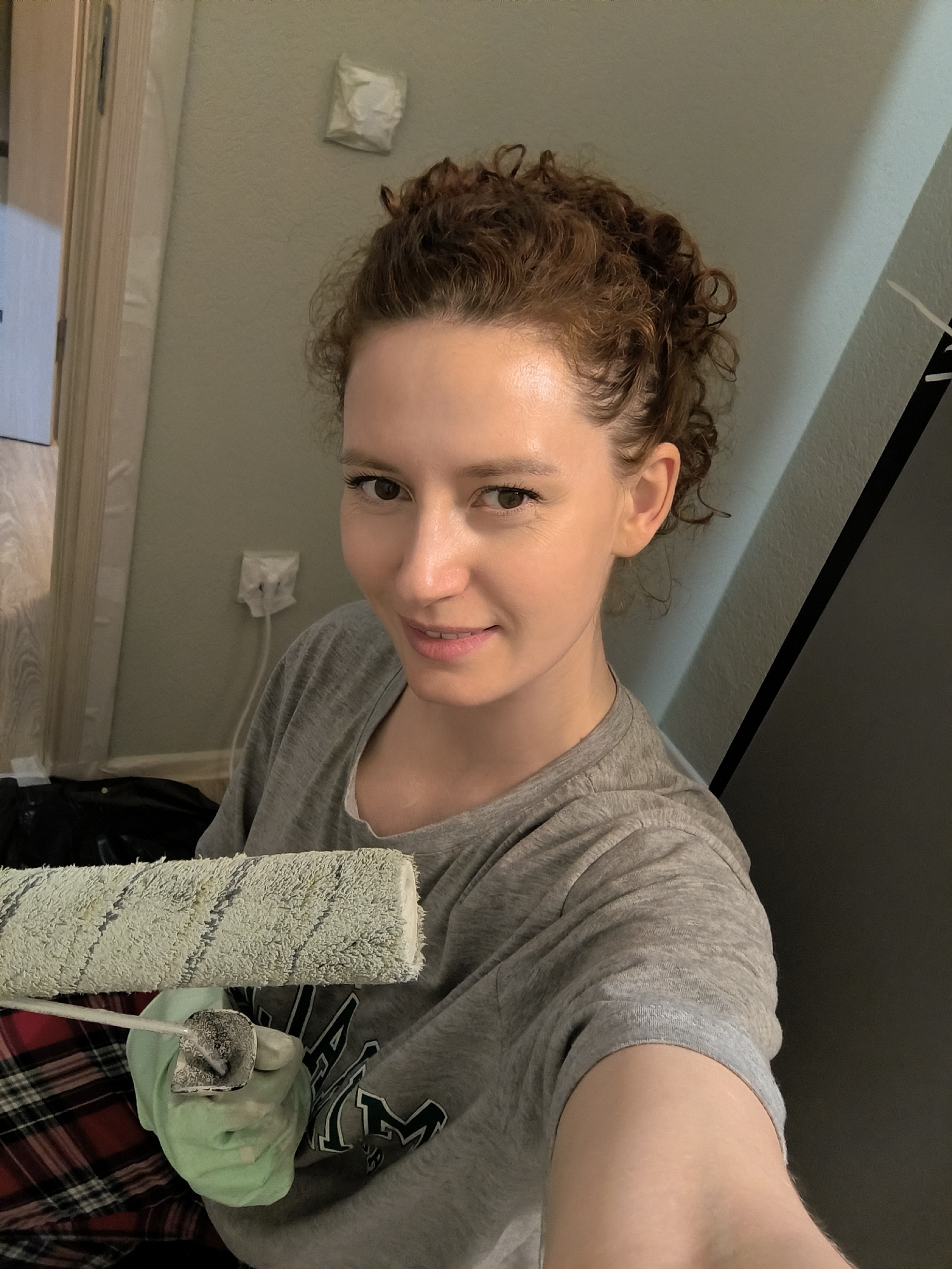 An indoor scene shows a woman taking a close-up selfie while engaged in painting. The subject, a female with fair skin and reddish-brown curly hair pulled back, occupies the majority of the frame, looking directly at the camera with a slight smile. She wears a grey t-shirt. In her left hand, covered by a light green glove, she holds a paint roller, its fuzzy surface appearing used and marked with light green and grey residue.
The background indicates an interior space undergoing renovation or painting. The wall directly behind her is painted a light grey-green hue. To the left, a light-colored wooden door frame is visible, partially covered with what appears to be protective tape or sheeting. A white electrical outlet with a white cord plugged into it is mounted on the wall. In the lower left quadrant, a red and black plaid fabric, possibly part of clothing or a drop cloth, is visible, along with a dark object resembling a garbage bag on the floor. To the right of the woman, a dark vertical surface, possibly a door or cabinet, is partially seen. The specific location in the city of Saint Petersburg, Russia, cannot be determined from the visual information provided.