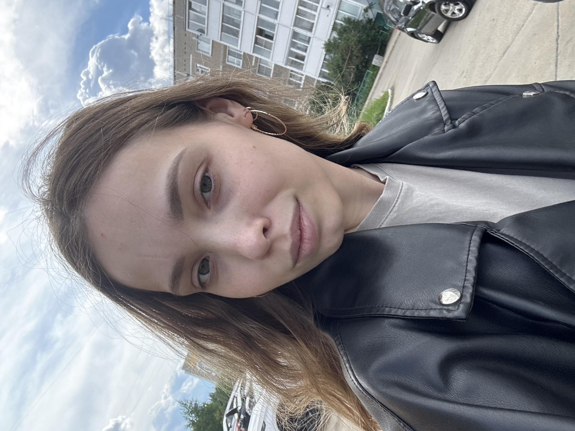 A young, light-skinned woman with brown hair is taking a selfie. She is wearing a black leather jacket, a light gray t-shirt, and gold hoop earrings. The background features a cloudy sky, a multi-story building with many windows, trees, and cars. The location appears to be in a residential area, which might be within the city of Chaikovsky, Russia.