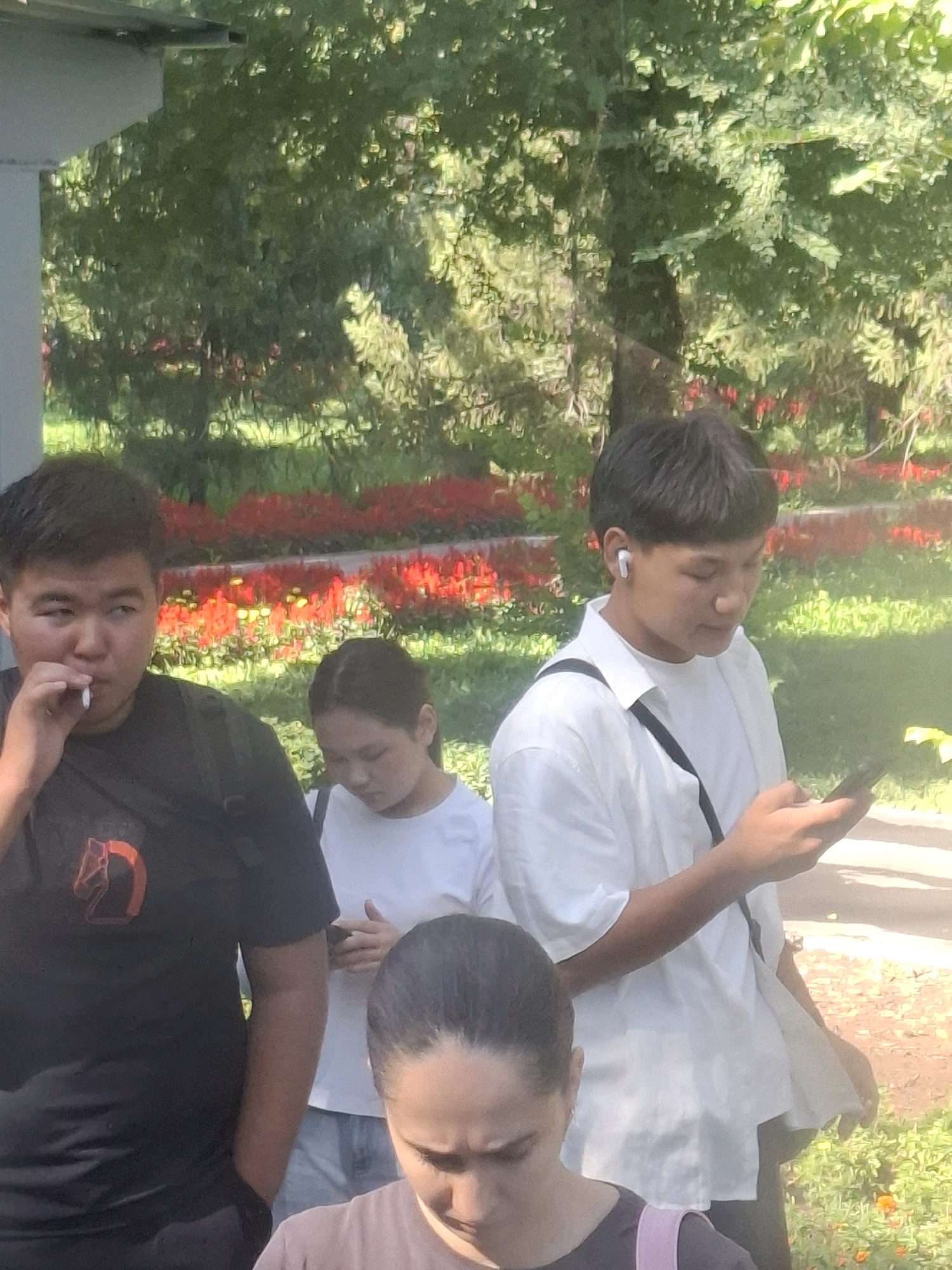 In Almaty, Kazakhstan, on a sunny day, four young people are gathered outdoors. In the foreground left, a young man wearing a black t-shirt with a red logo is smoking a cigarette, his left hand holding the cigarette and his right hand in his pocket. To his right, a young woman in a white t-shirt looks down. Partially visible behind her, a young woman in a pink top looks downwards. In the center right, a young man in a white shirt and earphones is looking at his phone, which he holds in his right hand. He has a black strap from a bag across his chest. The background is filled with lush green trees and a bed of red flowers, suggesting a park or garden setting. The ground is a mix of dirt and grass, with scattered small orange flowers visible on the right.