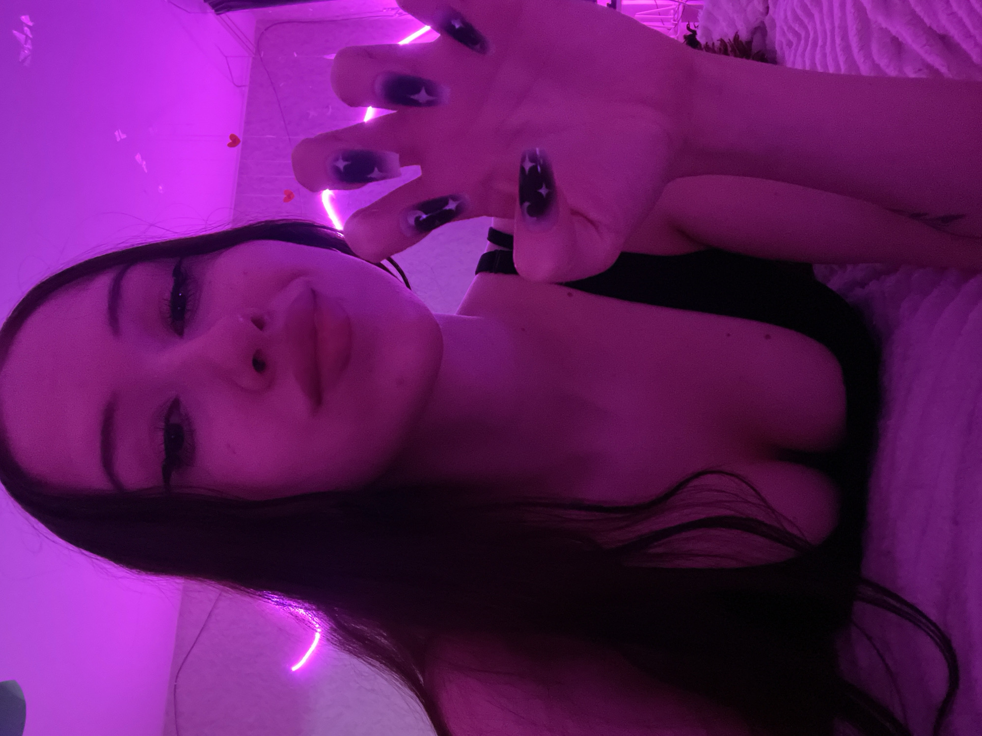 The image shows a young woman with dark hair and dark nail polish against a pink-lit background. She is wearing a black top and her right hand is in the foreground, with her fingers spread. The background appears to be a room with neon lighting and some small heart decorations. There are no discernible elements that would indicate a specific location in Votkinsk, Russia, or any other city.