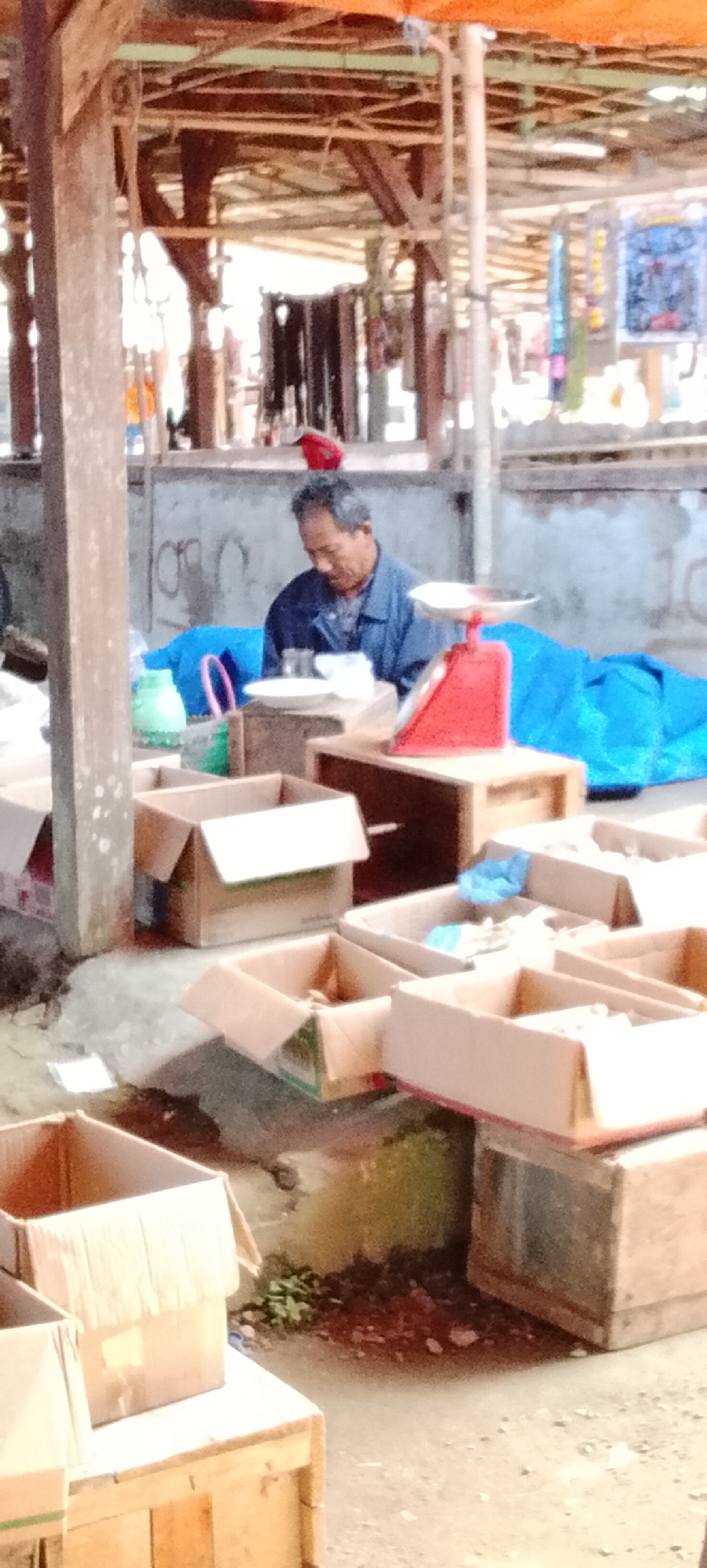 The image depicts an outdoor market stall in Bahway, Indonesia. A middle-aged man with graying hair, wearing a blue collared shirt, is seated behind a stall. In front of him are several open cardboard boxes, some appearing empty and others containing items that are not clearly discernible. A red analog scale is positioned on a wooden crate to the man's right, and a white plate is on another crate to his left. Behind the man, a large blue tarp is visible, and further back, various market stalls with hanging items and signage are present. The ground is a mixture of concrete and dirt, with some scattered debris and a small pile of green vegetation. The overall impression is one of a typical marketplace scene.