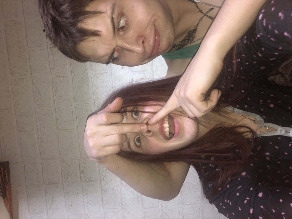 The image captures a humorous and playful moment between two individuals, presented at a 90-degree counter-clockwise rotation from the typical upright orientation.
In the upper part of the frame, one person, likely a young adult, has short, messy dark brown hair and light skin. They are wearing a light green tank top. A tattoo resembling a stylized feather or arrow design is visible on their upper chest/shoulder area. Their facial expression is exaggerated and playful, with eyes wide and looking to their right (which is upwards in the current orientation) and their mouth slightly open in a smirk or grimace. Their right hand is resting on the shoulder or upper arm of the second person.
In the lower part of the frame, a second person, also appearing to be a young adult, has long, reddish-brown hair that falls around their face and shoulders. They are wearing a dark long-sleeved top with light-colored patterns or polka dots, possibly pajamas. Their face is contorted in a wide, exaggerated expression, with their mouth wide open, revealing their teeth, suggesting they are either laughing loudly or making a playful yell. Their eyes are wide and looking towards the viewer. With their left hand, they are using their index finger to push up their own nose, further distorting their face. Simultaneously, their right hand is raised, with the middle finger extended in a "flipping off" gesture, directed generally upwards or towards the camera, but clearly intended in jest given the overall lighthearted nature of the photo.
The background consists of a light-colored brick or textured wall. The lighting appears to be artificial, casting some soft shadows. The overall mood of the image is casual, silly, and filled with youthful humor. There is no visible text in the image.