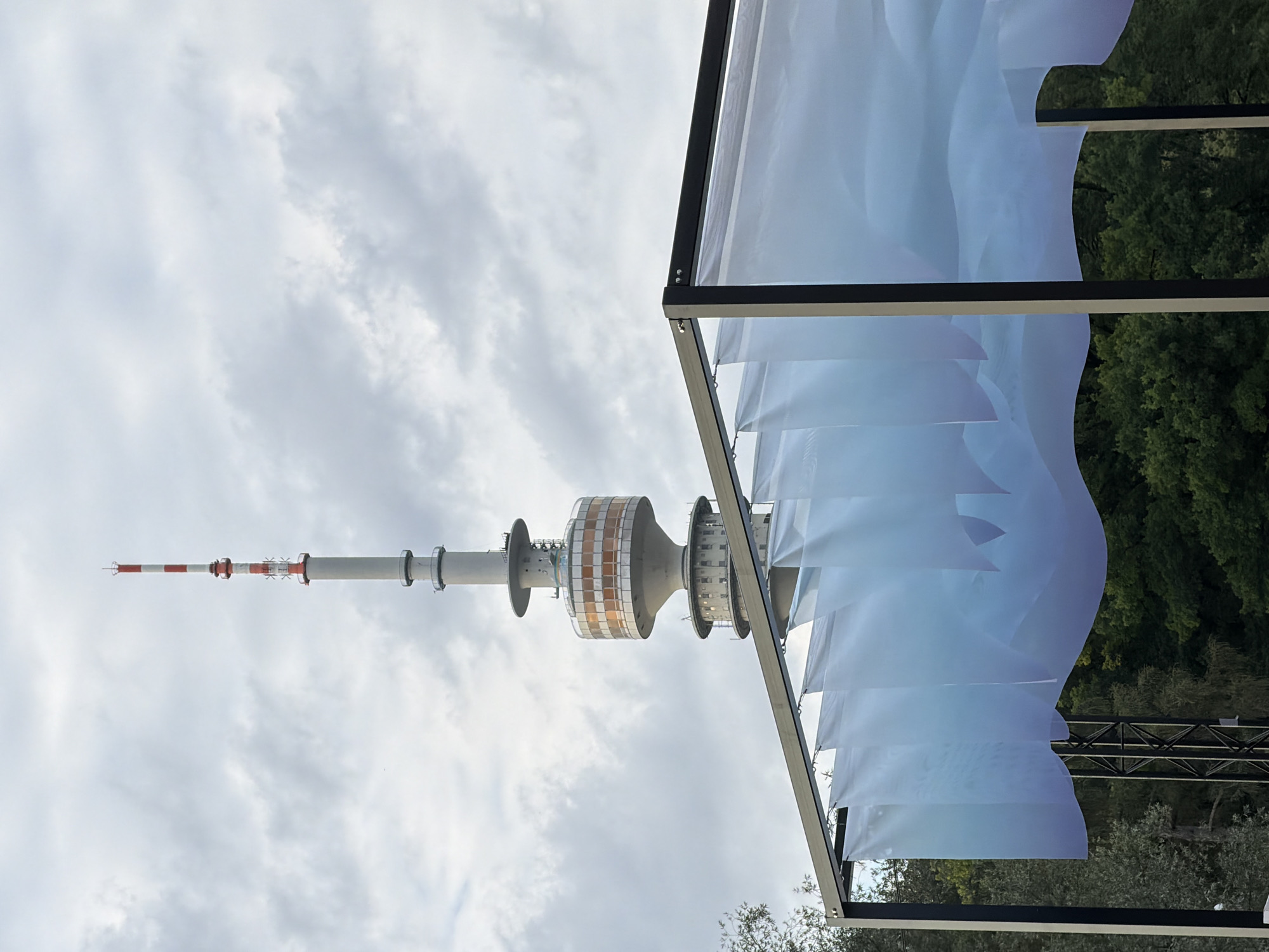 The image shows the Olympiaturm in Munich, Germany. In the foreground, there is a modern, open-air structure with a dark metal frame and light blue, translucent fabric panels hanging from the top. The fabric panels are draped and wavy, creating a sense of movement. Behind this structure, the Olympiaturm is visible. It is a tall, concrete tower with a red and white banded antenna at the very top. The tower has several levels, including a round observation deck with orange-tinted windows. The background consists of a cloudy sky and dense green trees. The scene appears to be set outdoors, possibly in a park or public space. There are no people visible in the image.