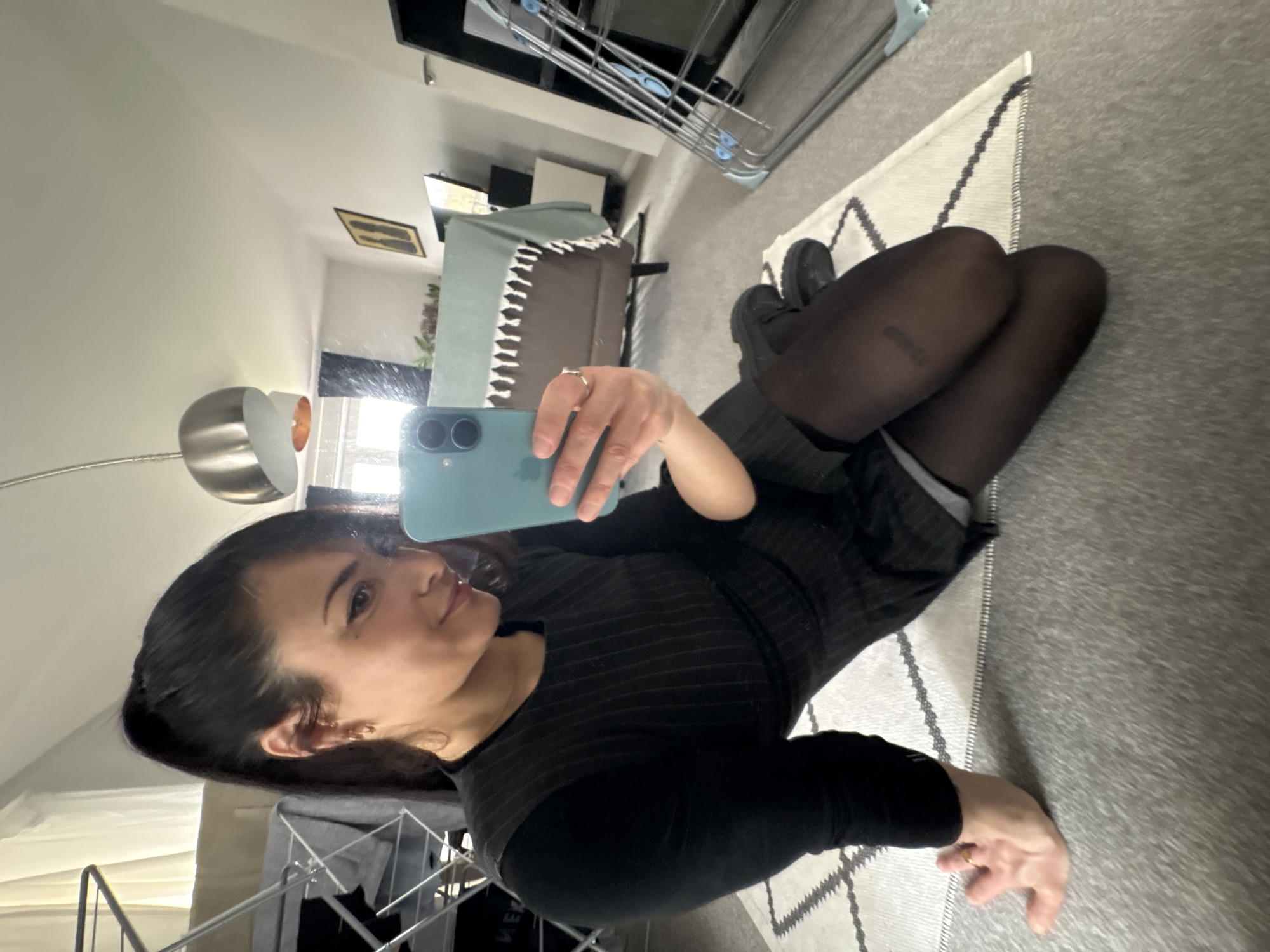 The image depicts a woman taking a selfie in a mirror. She is wearing a dark striped dress, black tights, and black shoes. She is holding a blue smartphone in her right hand. The background shows a room with a couch, a rug, and some furniture. The location cannot be determined from the image.