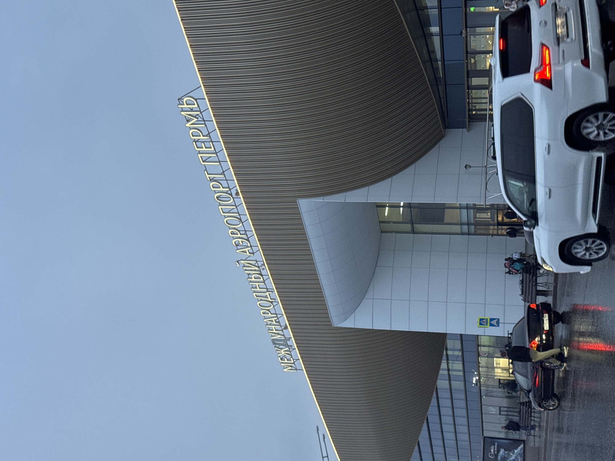 The image depicts the facade of the Perm International Airport in Bolshoye Savino, Russia, as indicated by the signage "МЕЖДУНАРОДНЫЙ АЭРОПОРТ ПЕРМЬ." The architecture features a curved, ribbed roof extending from the building. In the foreground, two cars are parked. The first is a white SUV, and the second is a black sedan with a person next to it. The ground is wet, reflecting the car lights. People are visible in the background near the building entrance.