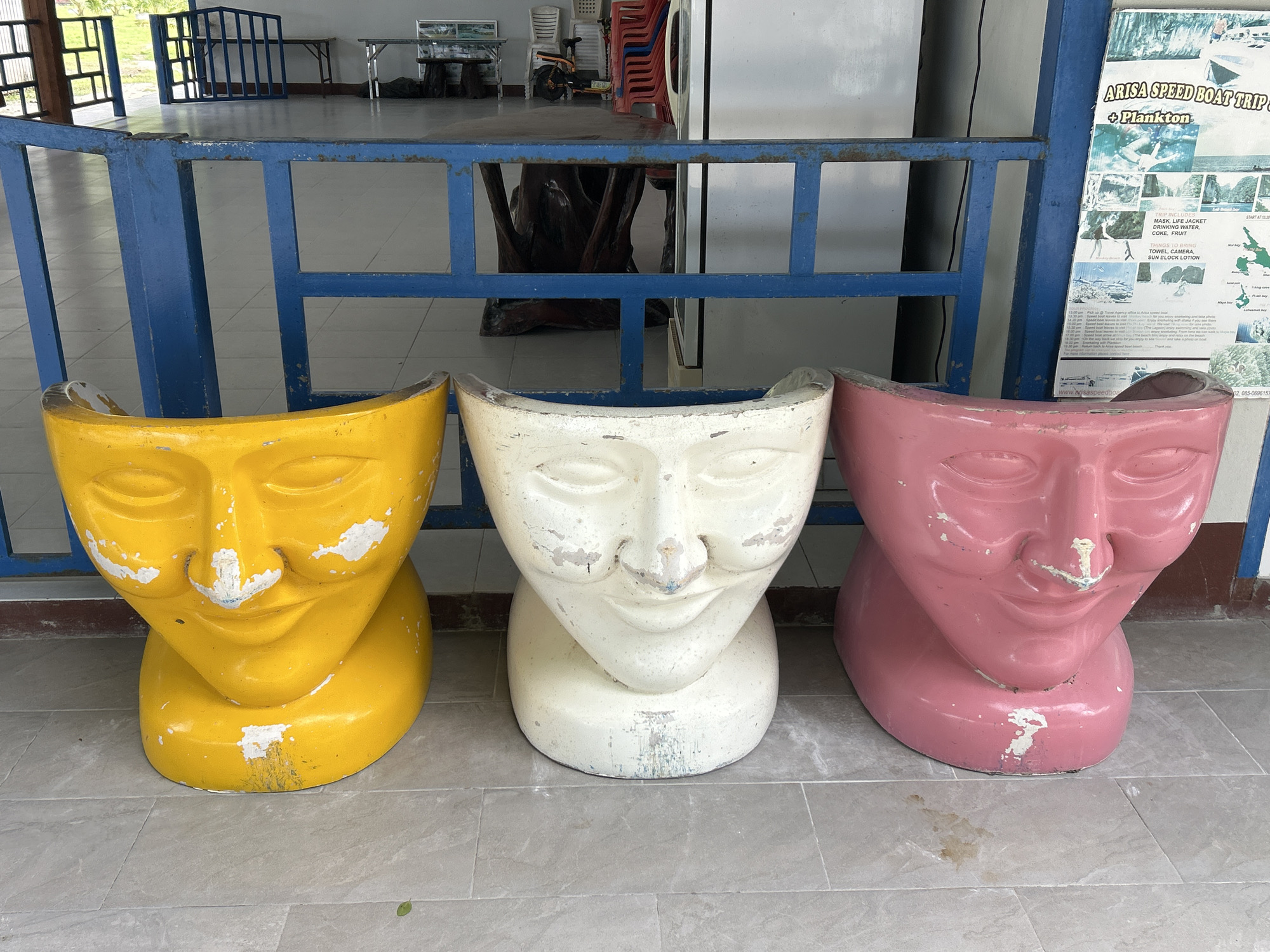 The image shows three large, stylized face sculptures, likely used as seats, positioned on a light gray tiled floor. From left to right, the sculptures are bright yellow, white, and pink. Each sculpture depicts an open-mouthed, closed-eyed face with a round base. The surfaces of the sculptures show signs of wear, with chips and scuff marks.
Behind the sculptures is a blue metal railing with square openings, and beyond that, a tiled floor and the interior of a building. In the background, tables, chairs, and a scooter are visible. To the far right, a poster titled "ARISA SPEED BOAT TRIP + PLANKTON" is affixed to a wall. The scene appears to be an outdoor waiting area or entrance, possibly at a pier or tourist information center in Ao Nang, Thailand, given the context of the poster.