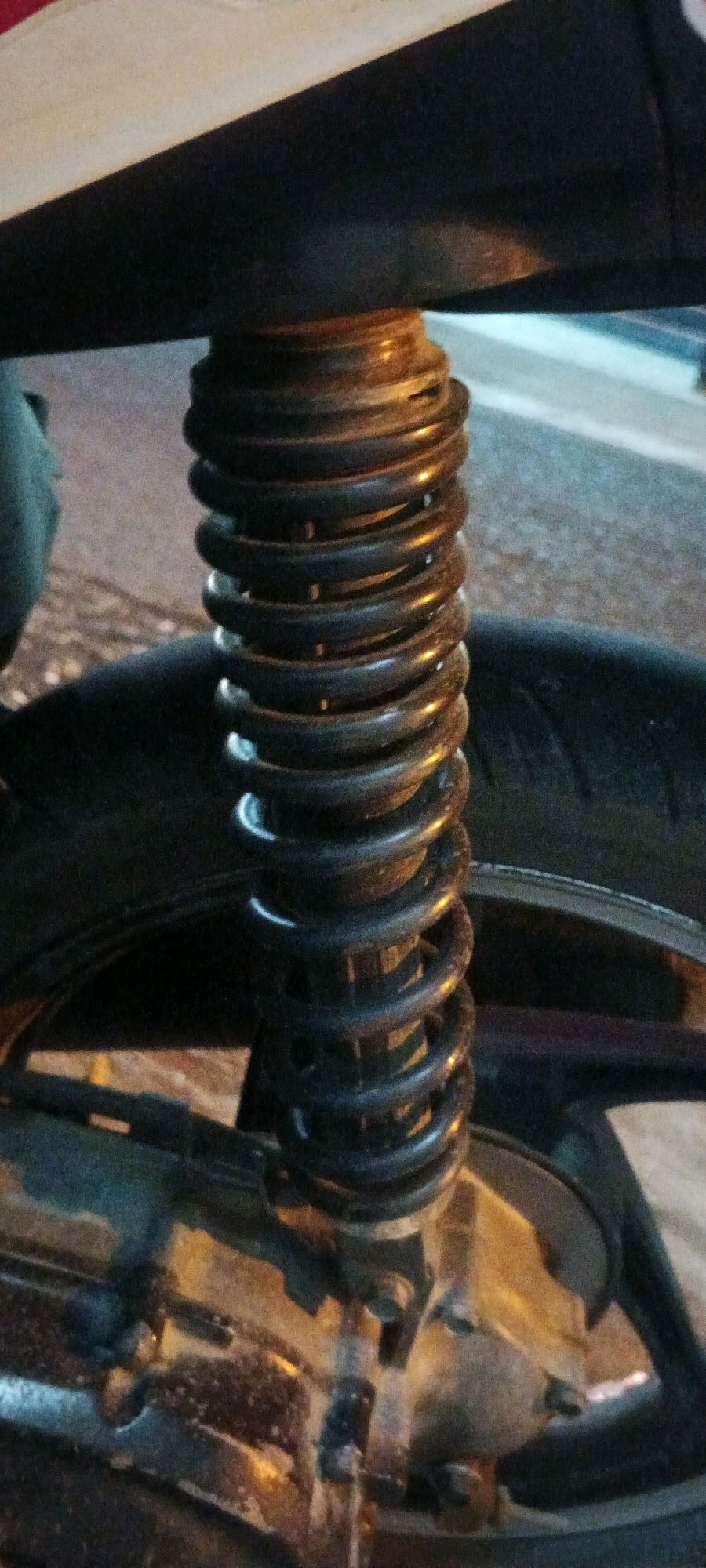 A close-up shot captures a black coil spring shock absorber on a motorcycle. The shock absorber is mounted vertically, with its top end secured to the motorcycle's body and its bottom end attached to the rear swingarm near the wheel. The spring is tightly wound, and its coils are visible. The background is blurred, but a motorcycle tire and part of the rear wheel can be seen. The scene appears to be outdoors, possibly on a street or parking area, with artificial lighting suggesting it may be evening or night. The location is identified as Kesiman, Indonesia.