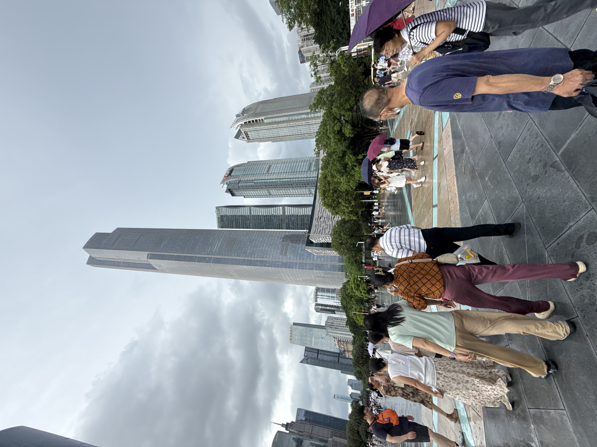 In Tianhe District, China, a wide, paved plaza is filled with a crowd of people walking under a cloudy sky. The plaza is surrounded by numerous modern skyscrapers of varying heights and architectural styles. The ground appears wet, suggesting recent rain, and some individuals are holding umbrellas. People are dressed in casual summer attire. In the foreground, a man in a blue t-shirt and a woman in a striped shirt are walking away from the camera. Further back, a group of women are walking together, some with umbrellas. The overall atmosphere is busy and urban.