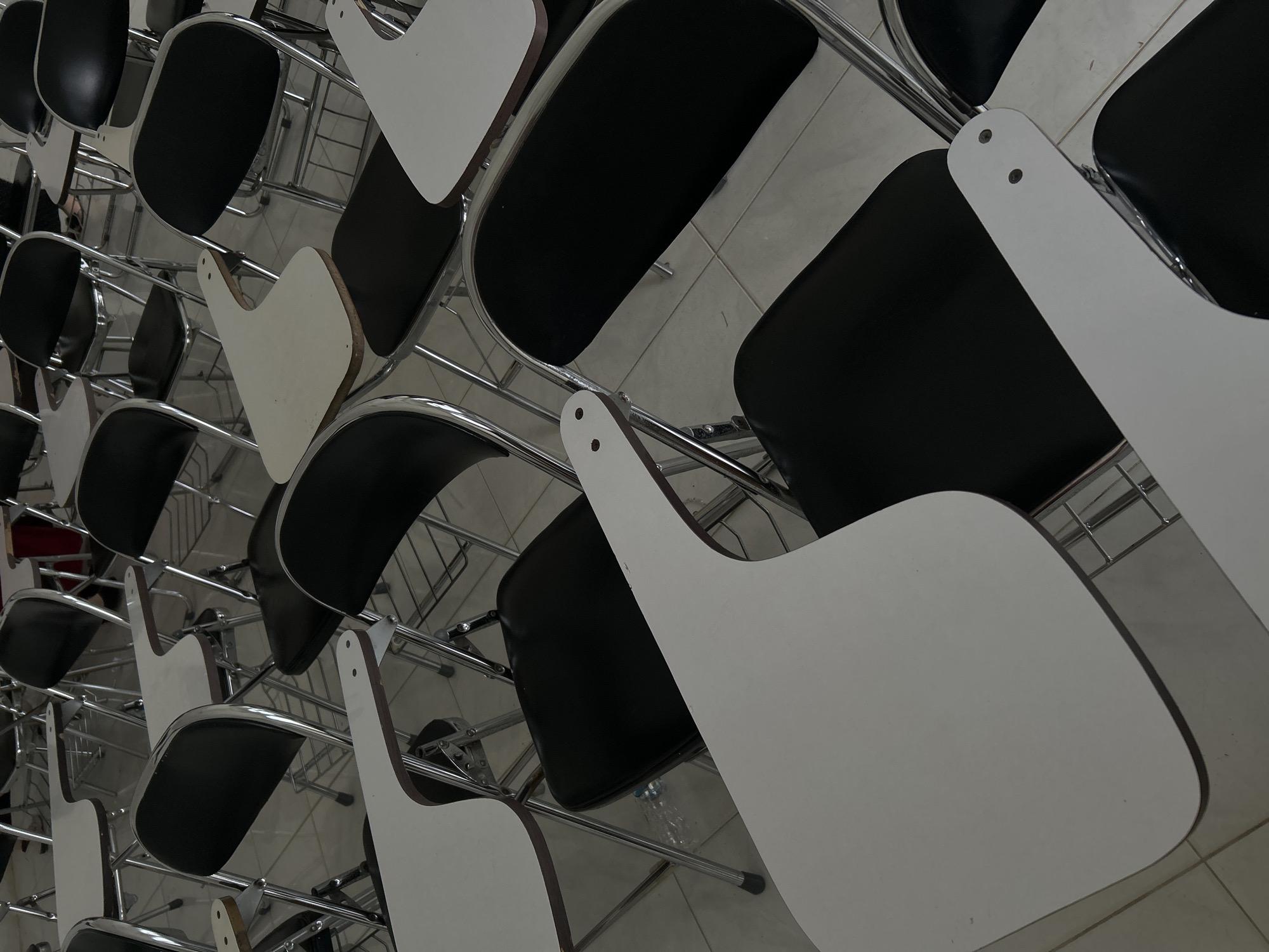The image captures an overhead, diagonal view of multiple empty chairs, likely situated in a classroom, lecture hall, or training facility in Reuleut Barat, Indonesia. The chairs are arranged in orderly rows, extending across the frame.
Each chair features a black, possibly upholstered, seat and backrest, supported by a shiny chrome metal frame. A key detail of these chairs is the white, rounded rectangular writing tablet attached to the right side of each chair, which pivots over the seat area. These tablets appear to have a dark edge, suggesting a laminate or wooden core. Small silver screws or fasteners are visible where the tablets connect to the chair frames.
The floor is made of light-colored tiles, possibly white or light grey, with visible grout lines forming a grid pattern. The lighting appears even and diffused, consistent with an indoor environment, providing no strong shadows to indicate direct sunlight.
The scene depicts an unoccupied space; there are no people present, and no activity is occurring. The empty chairs convey a sense of quietness or anticipation, suggesting the room is either awaiting an event or has recently been vacated. The repetition of the chair design and arrangement creates a rhythmic pattern across the image. No visible text is present.