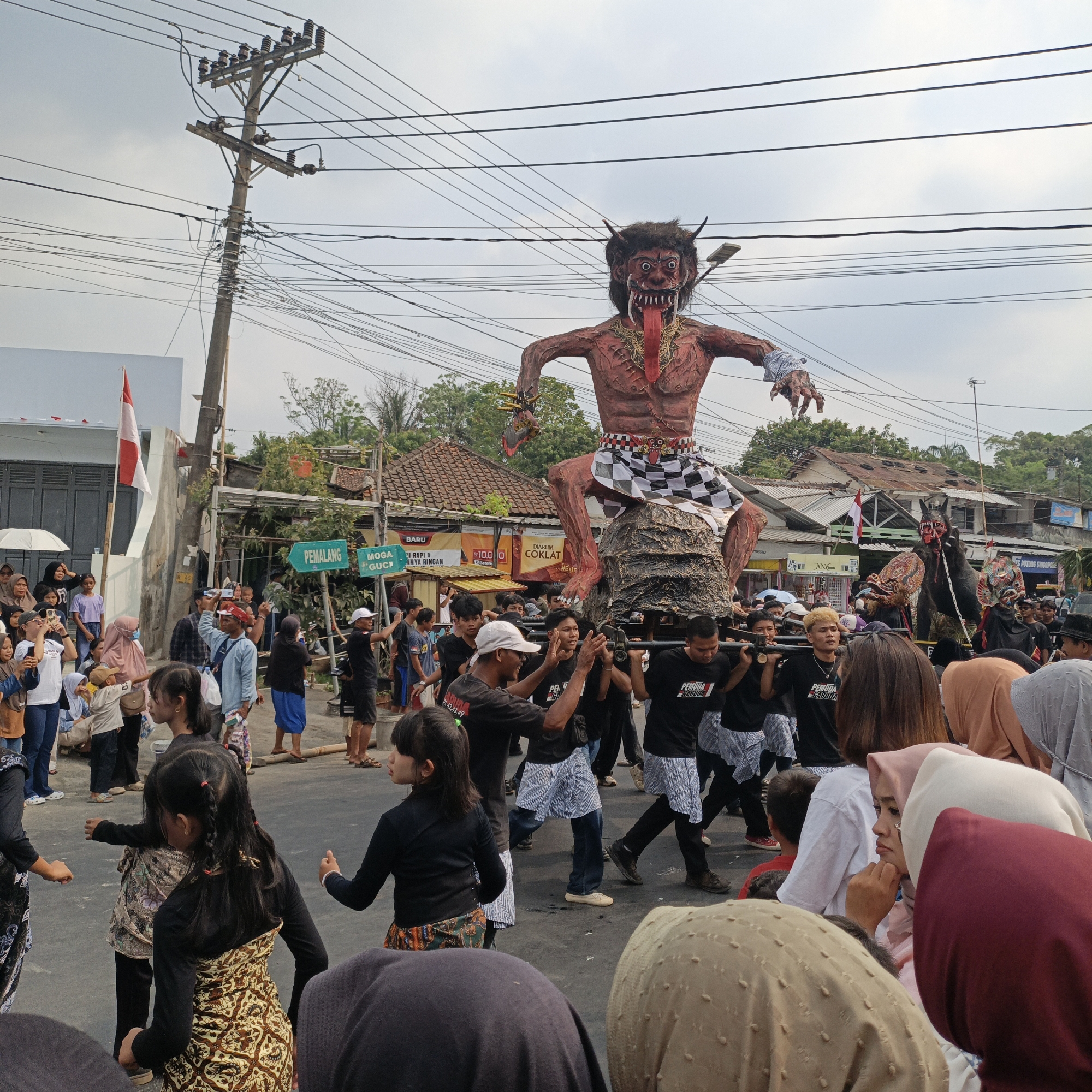 In Randudongkal, Indonesia, a large, intricately crafted effigy of a demonic figure, with a grotesque face, sharp teeth, and a protruding tongue, is being carried through a street. The effigy, predominantly red and brown, is adorned with a black and white checkered loincloth and sits atop a dark, rough texture base. It is supported by a group of men in black shirts and striped pants, who are marching in unison. Surrounding the effigy is a crowd of people, many of whom are observing the procession, with some holding up their phones to capture the event. Children are visible in the foreground, some dressed in traditional attire. The street is lined with buildings, shops, and utility poles with numerous wires crisscrossing the sky. A white and red Indonesian flag is displayed on a building in the background, and street signs indicating directions like "PEMALANG" and "MOGA GUCI" are also present. The sky is overcast, suggesting a cloudy day.