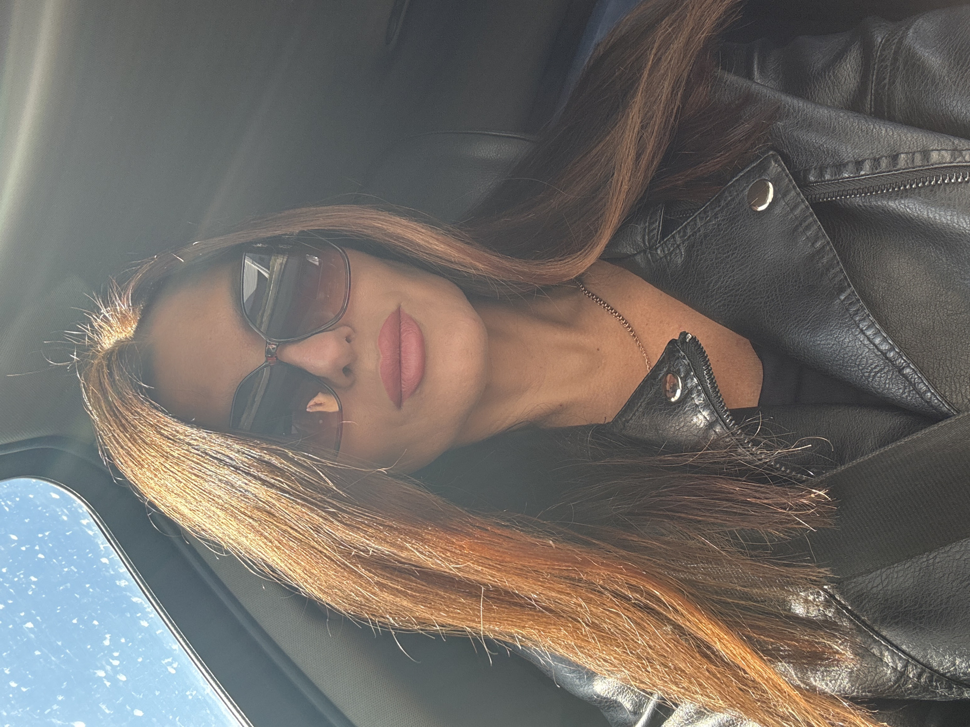 A woman is sitting in a car, wearing sunglasses and a black leather jacket. The image is angled, showing the woman from a slightly downward perspective. Part of the car's interior, including the sunroof, is visible. I cannot determine the location of the city of Chaikovsky, Russia from this image.