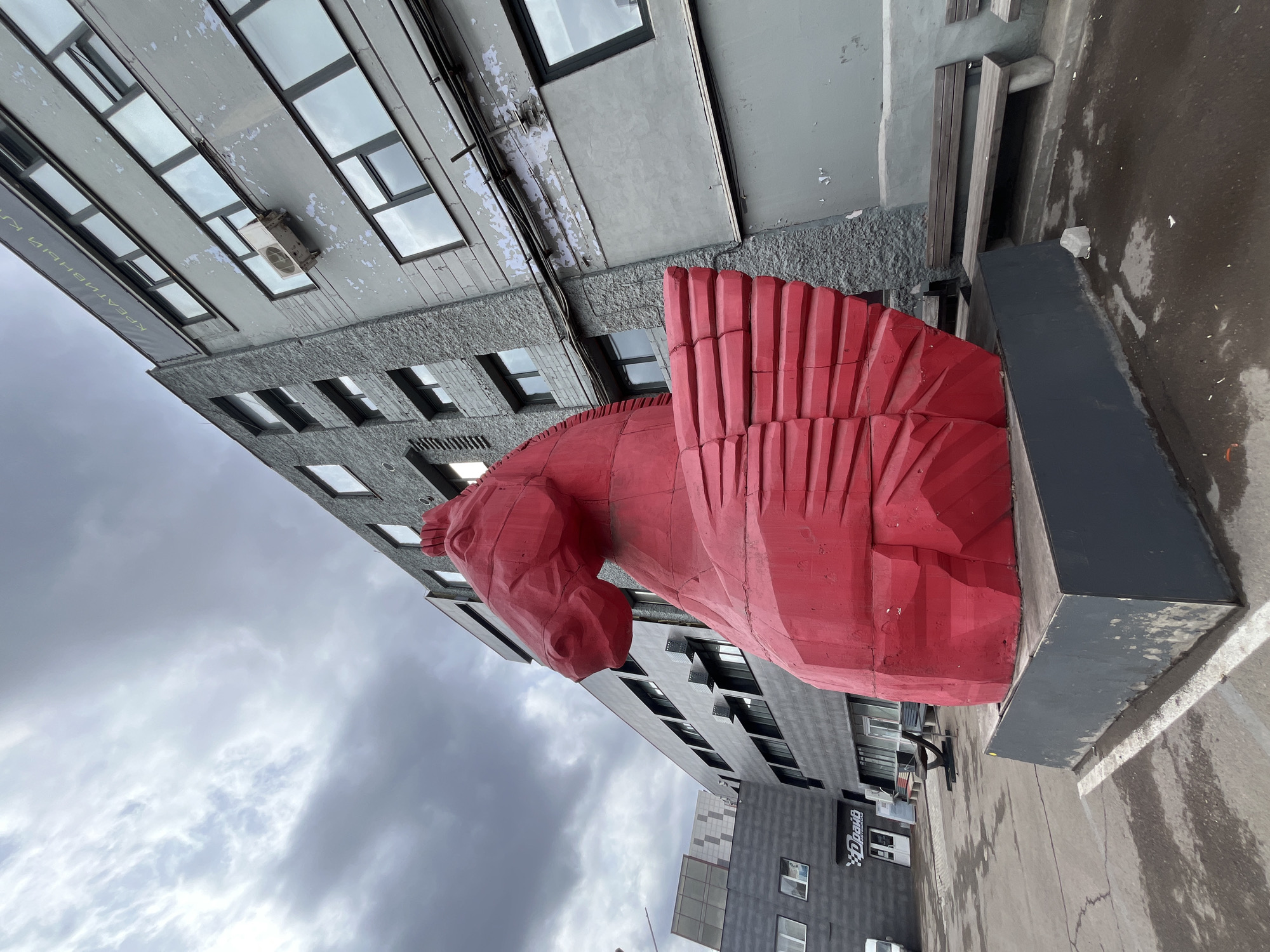 A large red sculpture of a horse with wings stands in front of buildings in Krasnoyarsk, Russia. The sky is overcast. The sculpture is on a gray base. The buildings are gray with windows. The street is wet.