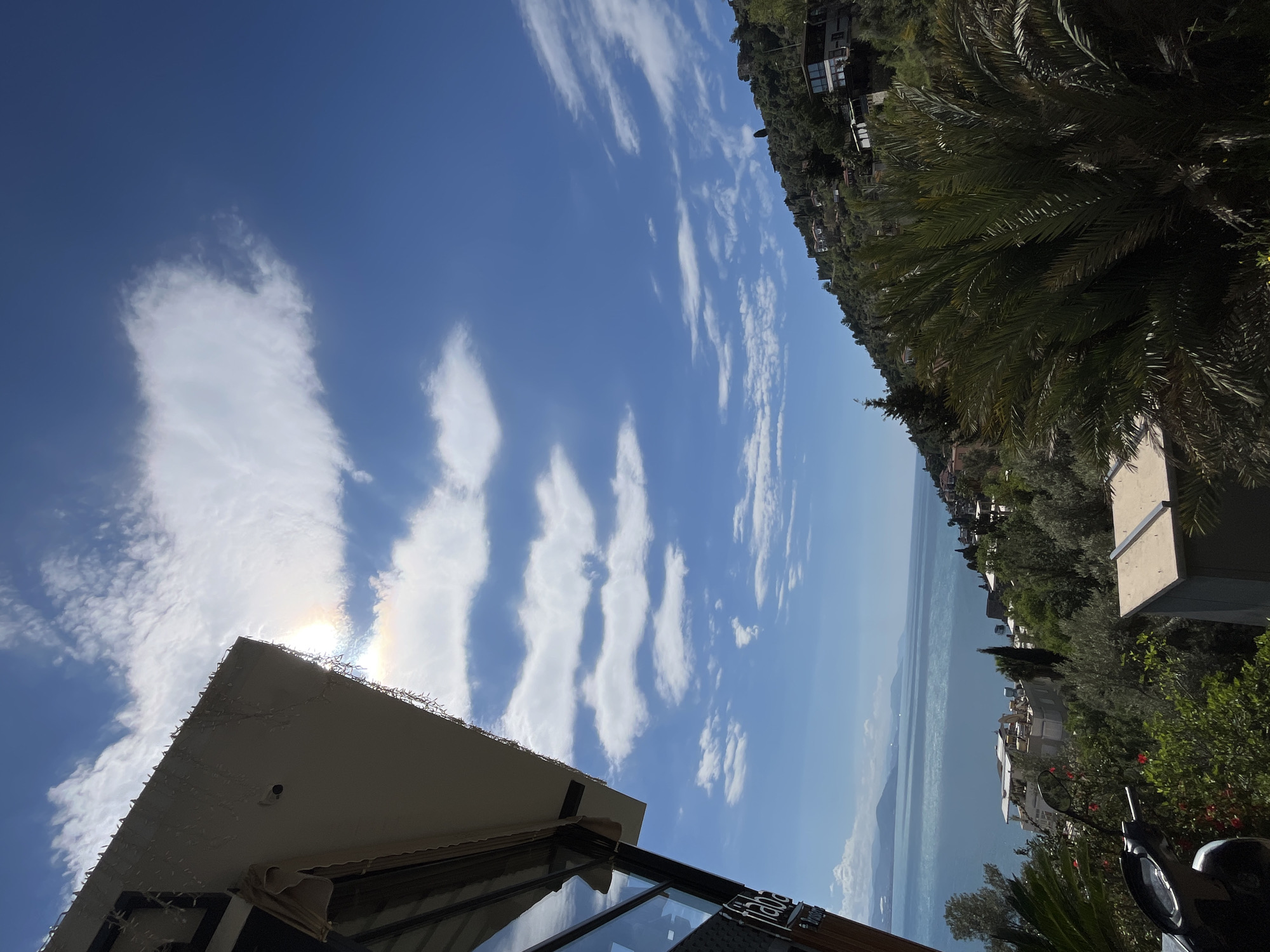 The image depicts a sunny outdoor scene in Alanya, Turkey. A clear blue sky with elongated cumulus clouds is visible. The sun is shining brightly, creating a halo effect around one of the clouds near the corner of a building.
The building, presumably a restaurant or café, has a white angled roof adorned with string lights along its edges. A glass window with a partial sign "Flaba Bar..." is seen below the roof.
In the background, there's a hillside with houses and trees, including a prominent palm tree. The blue sea is visible between the building and the hillside. In the foreground, there are some plants and a motorcycle.