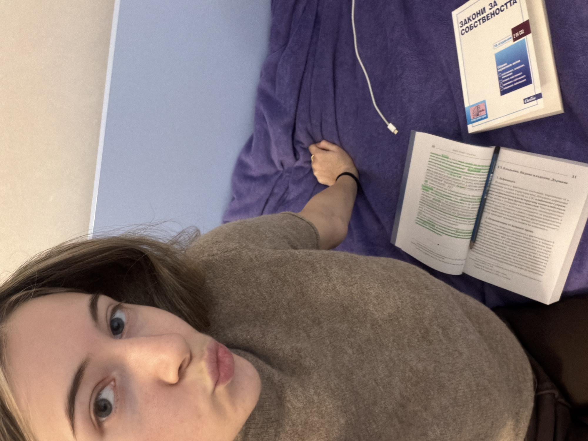 The image is a selfie taken from a high angle, looking down at a young woman lying down, with the camera rotated counter-clockwise. The woman, in the foreground, has light skin, long brown hair, and blue-grey eyes. She is looking directly into the camera with a "pouty" or "duck face" expression. She is wearing a textured light brown or beige knitted sweater. Her right arm is bent at the elbow, and a black elastic hair tie is visible on her wrist, along with a ring on one of her fingers.
The woman is lying on a light blue surface, possibly a pillow or headboard, while her arm and the objects beside her rest on a textured purple blanket or duvet. Two books are visible on the purple surface. One book is closed, with a white cover, blue and red graphic elements. The title on its cover, written in Cyrillic, is "ЗАКОНИ ЗА СОБСТВЕНОСТТА" (Laws on Property). Other text on this cover includes "VII. Изменение и допълнение" (VII. Amendment and Supplement) and "2-48 СО2," along with what appears to be a publisher's logo, "Ciela."
Next to the closed book, an open textbook lies face-up, with its pages displaying text. Some of the text on the left page is highlighted in green. Page numbers "30" and "31" are visible at the top corners of the open pages. A heading on page 31 is partially visible, reading "1. Владение, владение и държание. Характе...", which translates to concepts like "Possession, ownership and holding. Characte..." (likely characteristics). This content aligns with the "Laws on Property" theme of the closed book. A white charging cable with a USB connector is also visible on the purple blanket, near the books. Part of a light beige wall or surface is visible in the upper left background. The overall scene suggests the woman is taking a break while studying.
