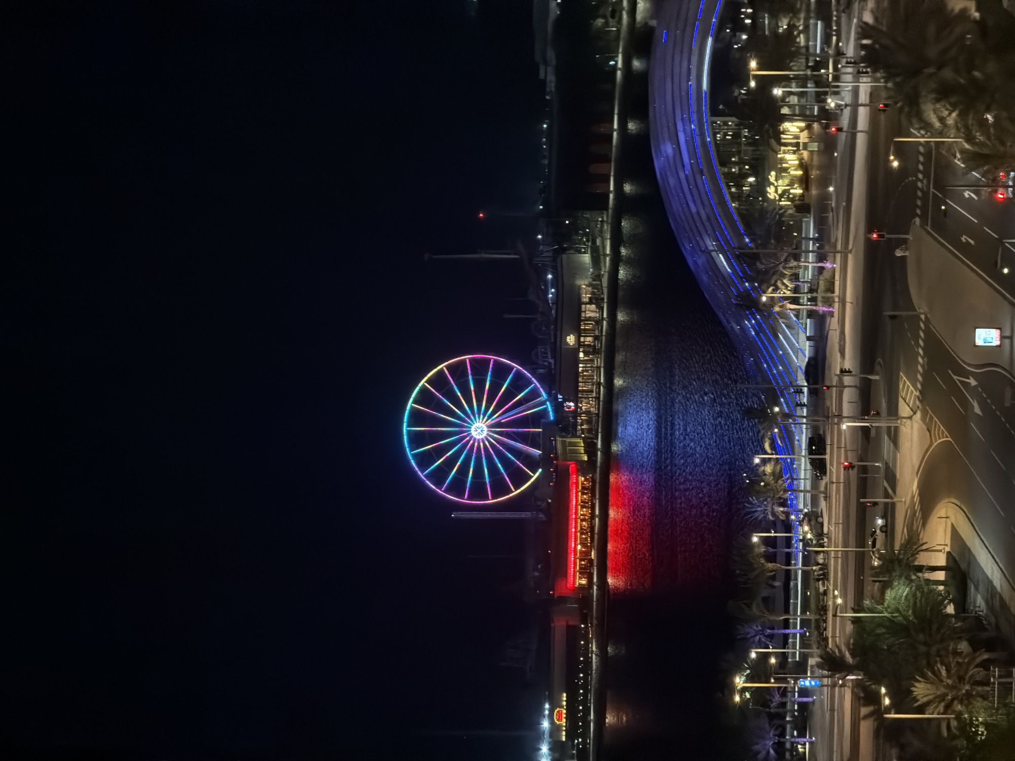 This is an elevated nighttime view of the Yas Bay Waterfront on Yas Island, Abu Dhabi, United Arab Emirates. The scene is dominated by a large, multicolored Ferris wheel in the background, contrasting against a dark sky.
In the foreground, a multi-lane asphalt road system curves through the frame, marked with white lines for lanes and pedestrian crossings. Several tall streetlights illuminate the road and adjacent sidewalks. Traffic lights along the road display red signals. Numerous palm trees line the roads and sidewalks. A light-colored sedan is visible on one road segment, while indistinct groups of people walk on the sidewalks. A distinctive modern architectural structure, possibly a bridge or building canopy, with horizontal blue strip lighting, spans above a section of the road.
The midground features a body of dark water, likely a bay or canal, reflecting the vibrant lights from the surrounding buildings and the Ferris wheel. The reflections create prominent streaks of red and blue light on the water's surface. Along the waterfront, several modern commercial buildings are illuminated. One building prominently displays a red-lit facade, which casts a strong red reflection onto the water. An illuminated sign for "The Cheesecake Factory" is discernible on this building. Another building to the left has a yellowish illuminated sign.
The background is dominated by the large Ferris wheel, brilliantly illuminated with a spectrum of colors including pink, purple, blue, green, yellow, and orange lights radiating from its center and outlining its structure. Behind the Ferris wheel, several tall, dark industrial or ride structures are visible against the dark night sky, one of which has a small red light at its apex. Distant scattered lights mark the horizon. The overall impression is that of a lively entertainment and commercial district at night.