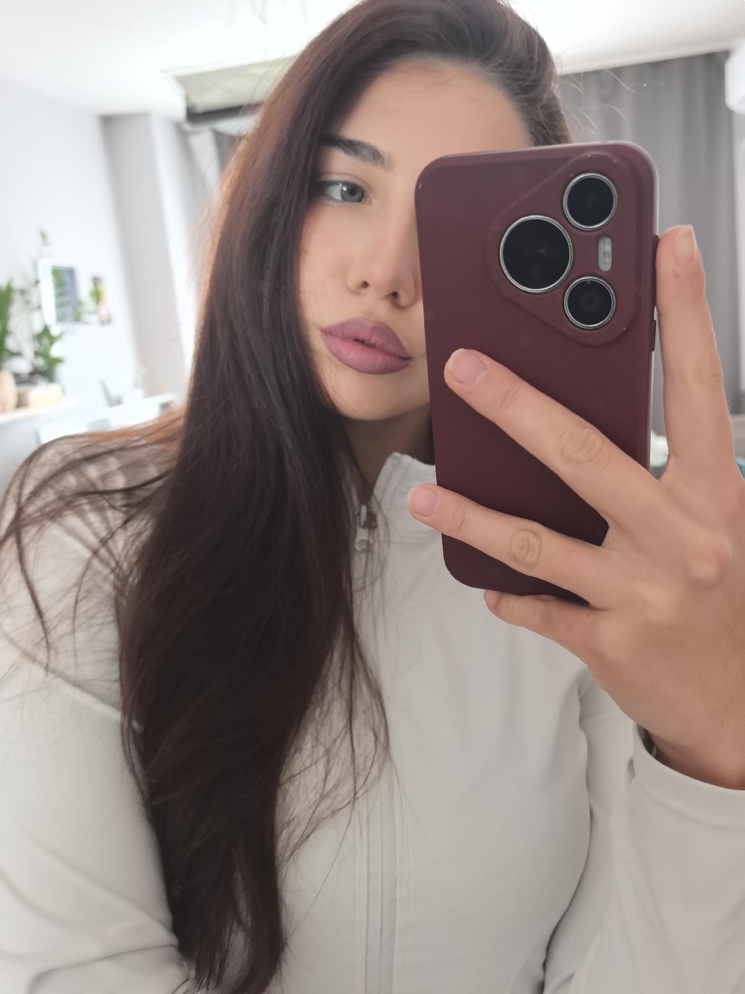 A young woman is captured in a mirror selfie, focusing on her upper body and face. Her long, dark brown hair is prominent, flowing over her shoulders. She has light-colored eyes, possibly blue or green, and is wearing bright pink lipstick. She is dressed in a light-colored, long-sleeved top or jacket, possibly white, with a zipper detail visible at her neck.
Her right hand holds a smartphone encased in a maroon or burgundy protective cover. The back of the phone features a distinct camera module with three circular lenses. The setting appears to be an indoor room, characterized by light-colored walls. In the blurred background, reflections reveal elements such as a green plant in a pot on the left, and a ceiling-mounted unit (possibly an air conditioner) in the upper right. The image depicts a casual, daytime moment in what appears to be a residence in Stara Zagora, Bulgaria. No text is visible in the image.