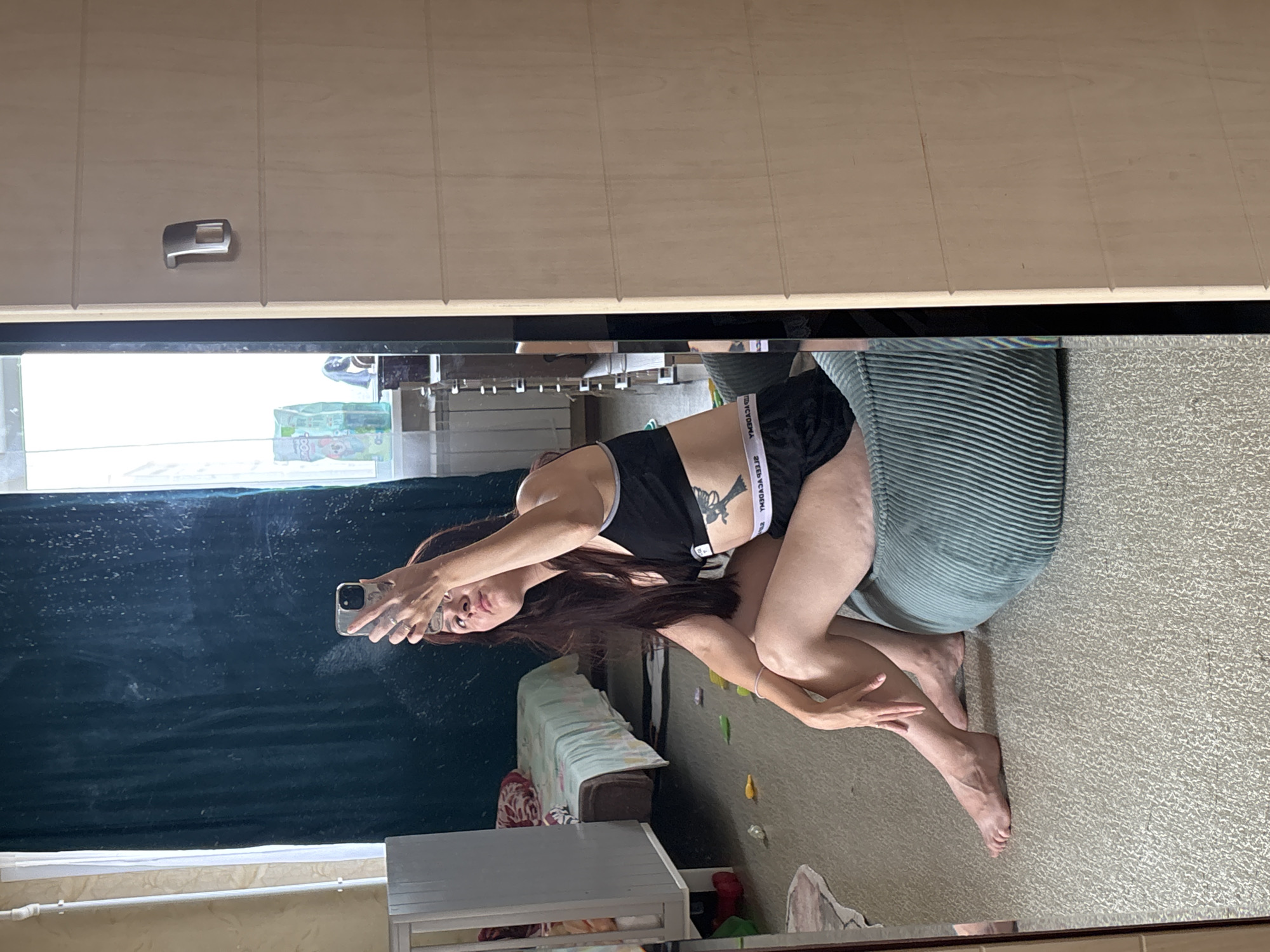 A woman is taking a mirror selfie in a room. She is sitting on a teal beanbag chair, wearing a black sports bra and black shorts. The room appears to be a bedroom, with a crib on the left side and a bed in the background. The floor is carpeted. The mirror reflects the woman and a portion of the room. The overall impression is a casual, indoor setting. The location of Naberezhnye Chelny, Russia, is not discernible from the image itself.