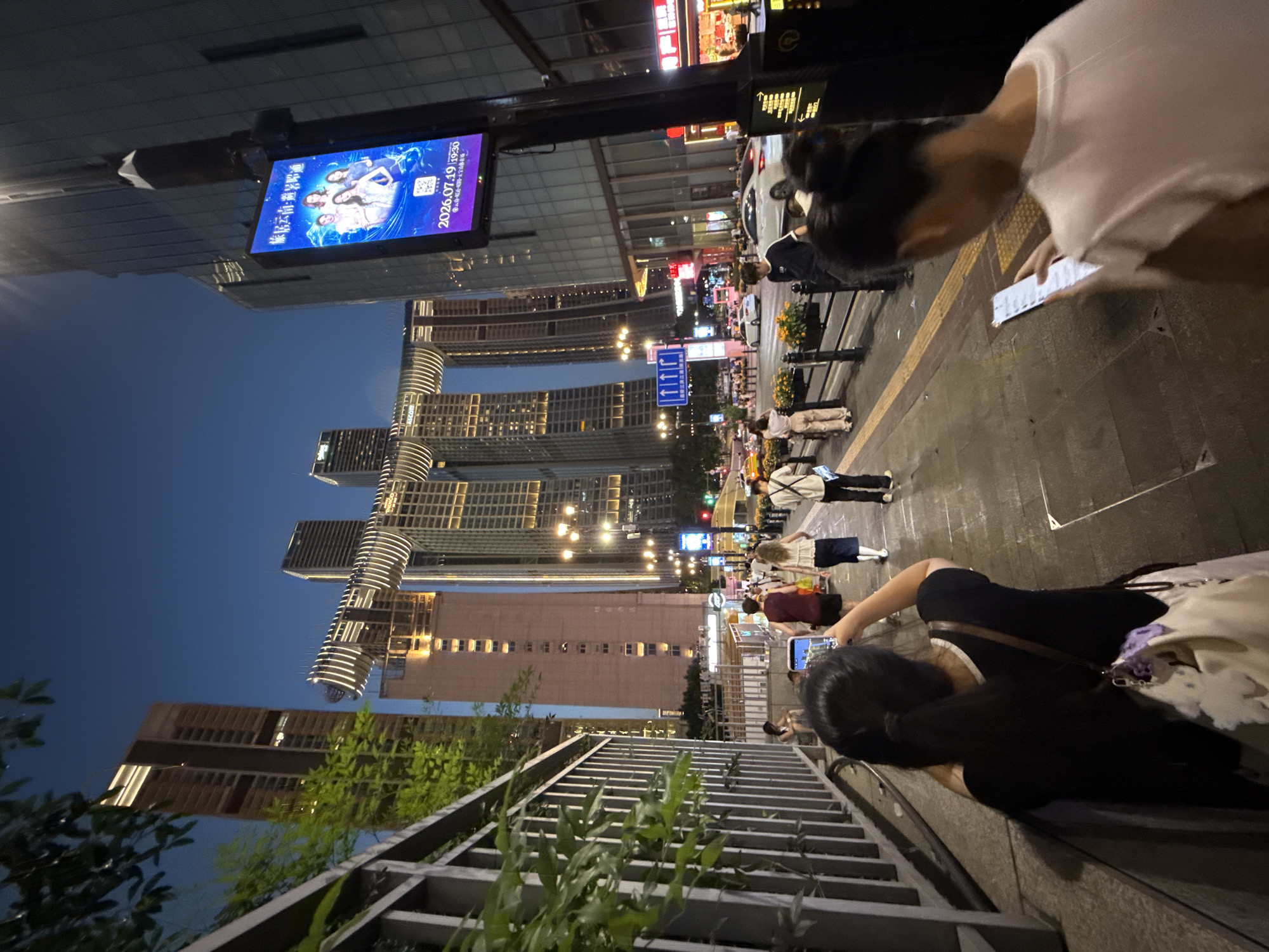 The image depicts a street scene at dusk in the Yuzhong District of Chongqing, China, likely near the Raffles City Chongqing complex, identifiable by the distinctive architecture of the interconnected buildings. In the foreground, two women are visible. The woman on the left, facing away from the camera, is wearing a black shirt and holding a smartphone up to take a picture. A light-colored shoulder bag with a purple charm hangs from her shoulder. The woman on the right, also facing away, is wearing a light pink shirt and looking down at her smartphone.
The middle ground shows a pedestrian walkway with several people walking. Some are dressed casually, while others appear to be tourists. Yellow tactile paving is present along the edge of the walkway. Beyond the walkway, a street with cars and a bus is visible. Traffic lights and streetlights illuminate the area.
The background is dominated by modern skyscrapers, including the iconic Raffles City Chongqing complex with its curved, interconnected bridge-like structures that are illuminated by white lights. The sky is a deep blue, characteristic of twilight. An electronic billboard displaying an advertisement with Chinese text and images of people is mounted on a pole to the right of the frame. A metal fence with green foliage runs along the left side of the image. The overall impression is an active urban environment at the transition from day to night.