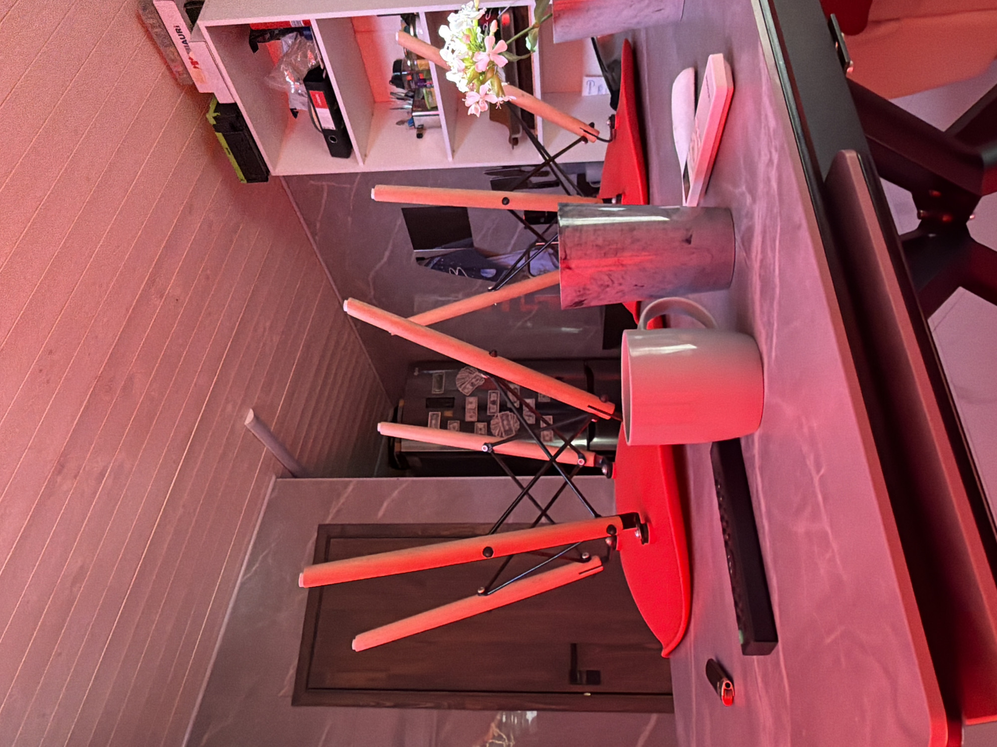 The image depicts an indoor scene, possibly a kitchen or dining area, illuminated by red lighting. The primary focus is a table on which several objects are placed. A black remote control and a small lighter are visible on the left side of the table. To the right of these, a light-colored mug sits next to a metallic-looking cup. Further to the right, a white object, possibly a remote control, rests on the table. Behind the table, the legs of several chairs are angled upwards, with one chair's red seat prominently displayed. In the background, a doorway is visible on the left, and a shelf unit is on the right, containing various items, including boxes and a bouquet of white flowers. The overall impression is one of disarray, possibly due to the chairs being upturned. There is no indication of the specific location in Orenburg, Russia, or any people present in the image.