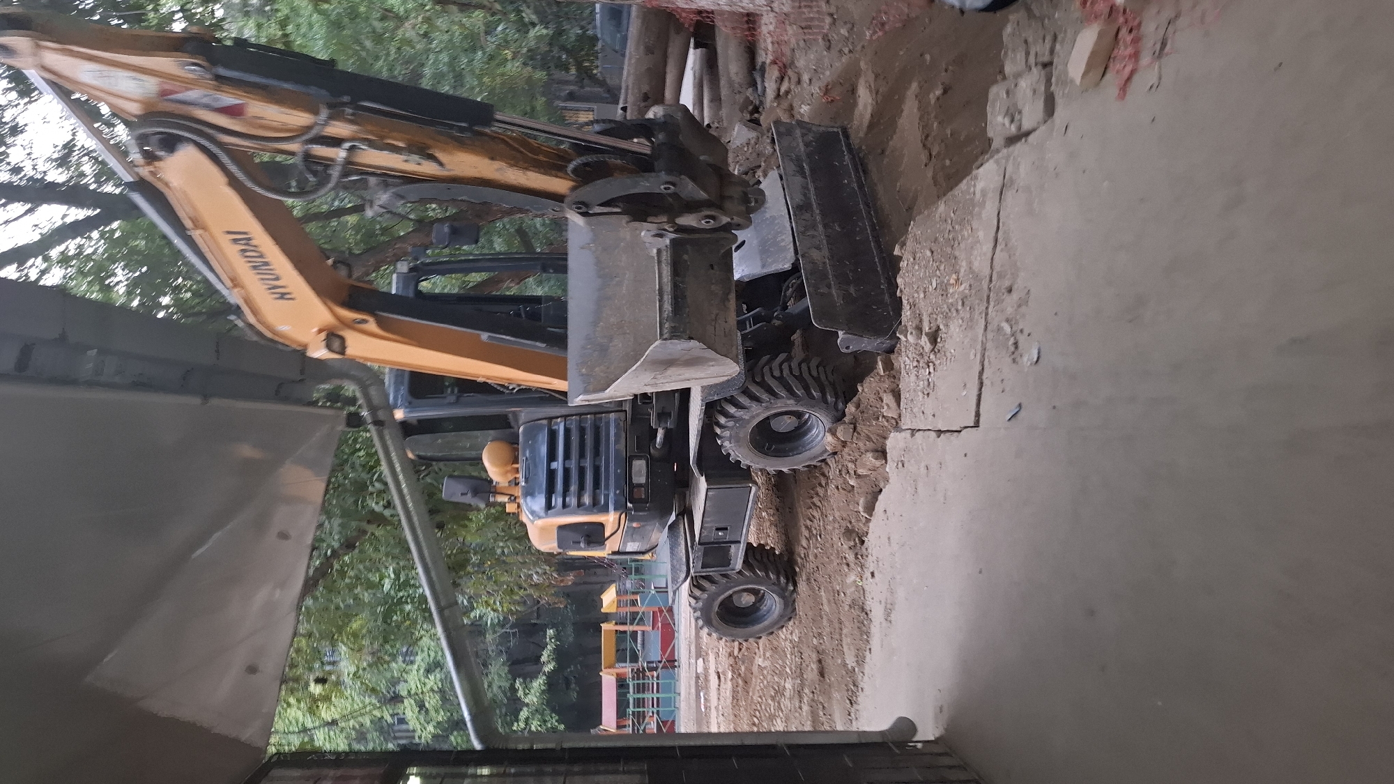 An orange and black Hyundai excavator is parked on a dirt construction site in Shymkent, Kazakhstan. The excavator is positioned diagonally in the frame, with its large bucket resting on the ground. Its tires are thick and rugged, suitable for rough terrain. In the background, there are trees and a partially visible playground with colorful structures. The foreground shows a concrete path and more dirt. The lighting suggests it is daytime, possibly overcast or late afternoon, as the light is diffused. No people are visible in the image. The scene depicts an active construction area in an urban setting.