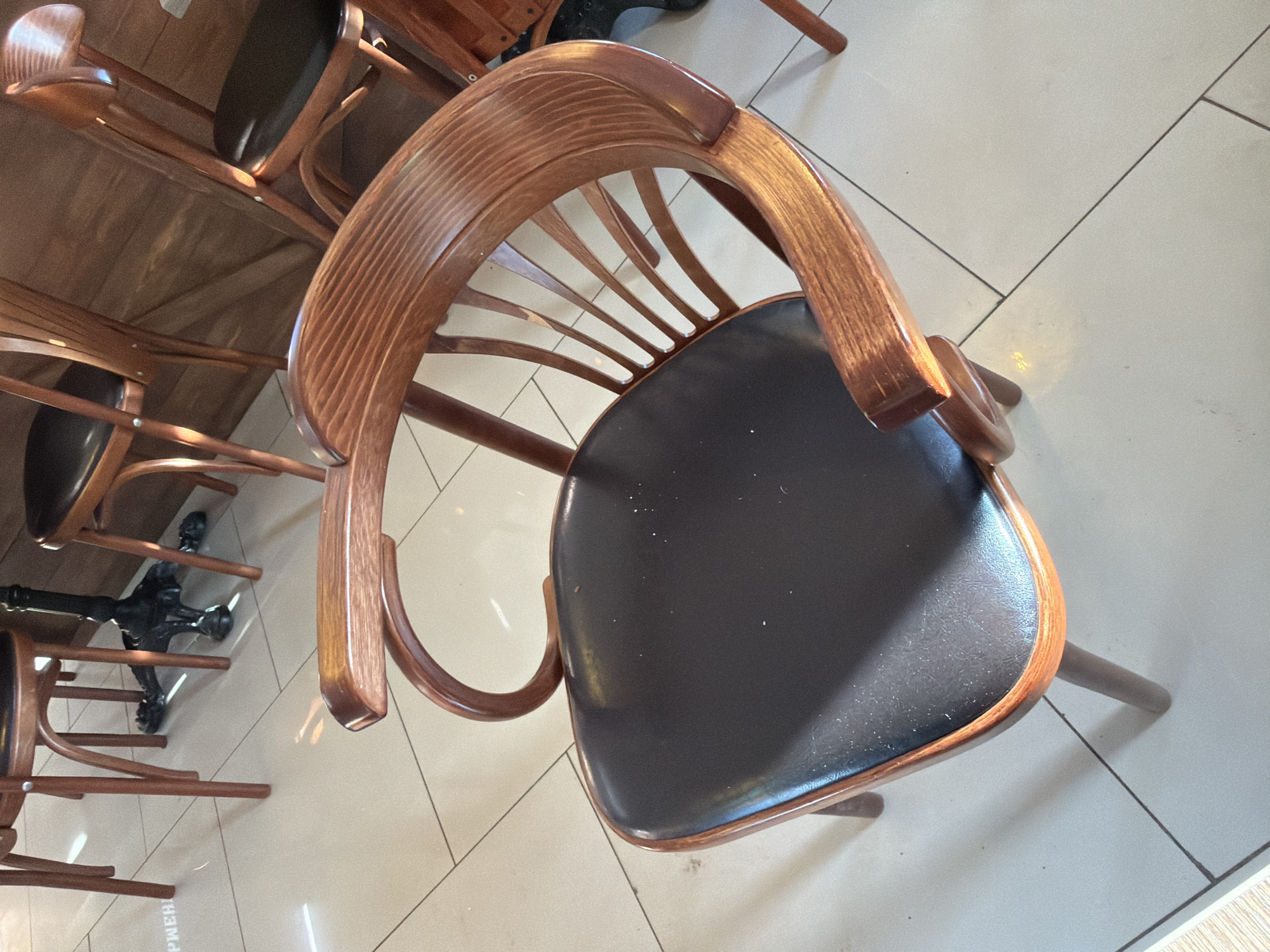 The image depicts a close-up, high-angle shot of a wooden chair with a dark seat. The chair is positioned on a light-colored tiled floor with visible grout lines. The chair is part of a set arranged around a table, with other chairs and the table visible in the background. The focus is on the single chair, highlighting its curved back and the texture of the wood. The scene appears to be indoors, possibly in a cafe or restaurant. There is no evidence of people or any identifiable characteristics to suggest that it is in Sarapul, Russia. The description is purely based on the visual elements present in the image.