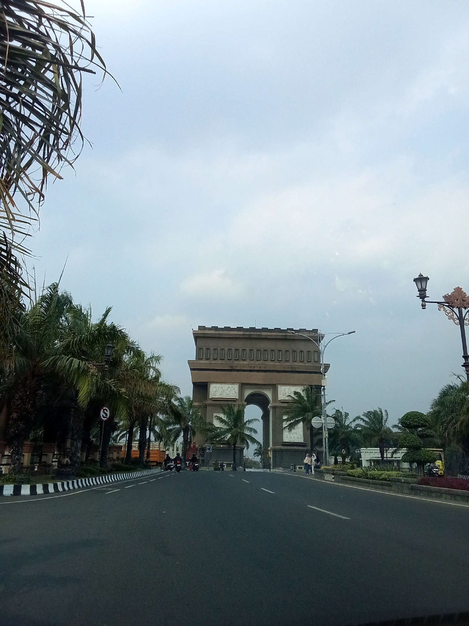 The image shows a street scene in Ngasem, Indonesia, dominated by a large, monumental archway. The archway, constructed of brown stone, features a prominent central arch and decorative elements along its top. It is flanked by palm trees on either side, which are also visible along the road. A street lamp with an ornate design stands to the right of the archway. The road in the foreground is asphalt, with a white and black striped curb. Several motorcycles are visible on the road, suggesting traffic. Two people are walking on the sidewalk near the archway. The sky is overcast, with some clouds visible. The overall impression is that of a public space in a tropical urban environment.
