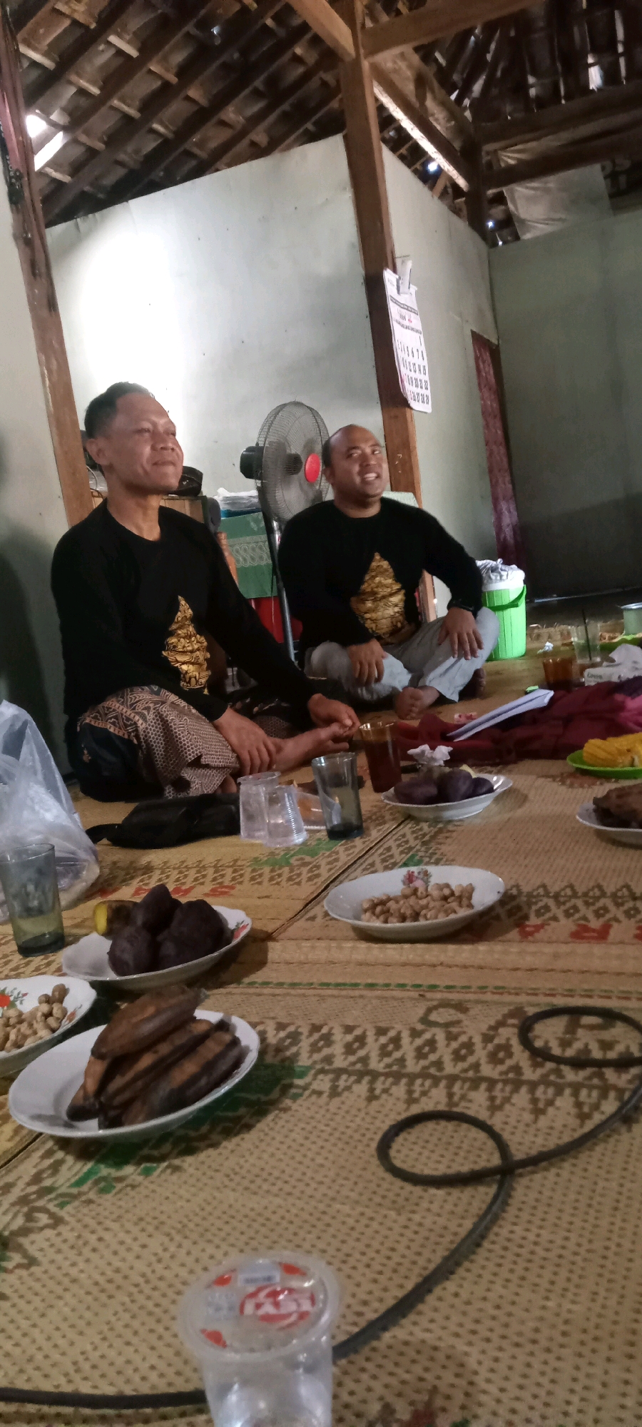 The image depicts an indoor scene, likely a dwelling, with two men seated on the floor on a woven mat. The man on the left is wearing a black long-sleeved shirt with gold intricate patterns and a patterned sarong. He has short, dark hair and is looking to his right. The man on the right is also wearing a black long-sleeved shirt with similar gold patterns and light-colored pants. He has a shaved head and is looking upwards and to his left, with his mouth open as if speaking. They are both barefoot.
In the background, a standing fan is visible. A calendar is attached to the wall, which is a light green color. The ceiling is made of wooden beams and what appears to be corrugated metal or similar material. There are various plates of food and drinks on the mat in front of the men. These include what appear to be bananas, small dark fruits, and nuts. Several glasses and cups are also present. A green plastic cooler is near the fan. The overall environment suggests a casual gathering or meal. The image is undated and unlocated, but the style of the house and the presence of the calendar with Indonesian script (visible in the zoomed-in crops) suggest a location in Indonesia. Without further information or visible landmarks, it is not possible to pinpoint the exact location within the city of Jering, Indonesia.