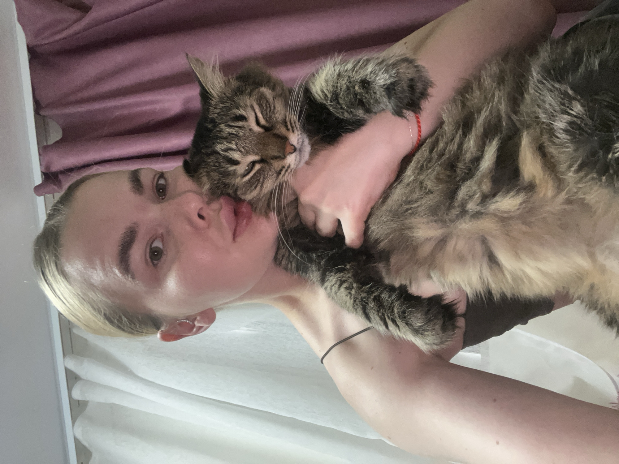 A young woman with blonde hair pulled back in a bun is holding a fluffy tabby cat. She is looking at the camera with her eyes partially closed and her lips pursed. She is wearing a dark-colored spaghetti strap top. The cat is nestled in her arms, and its eyes are also closed. Behind them, a sheer white curtain is visible on the left, and a pinkish-purple curtain is on the right. The image appears to be taken indoors, possibly in a bedroom. There is no specific information in the image to indicate the location is Tikhoretsk, Russia.