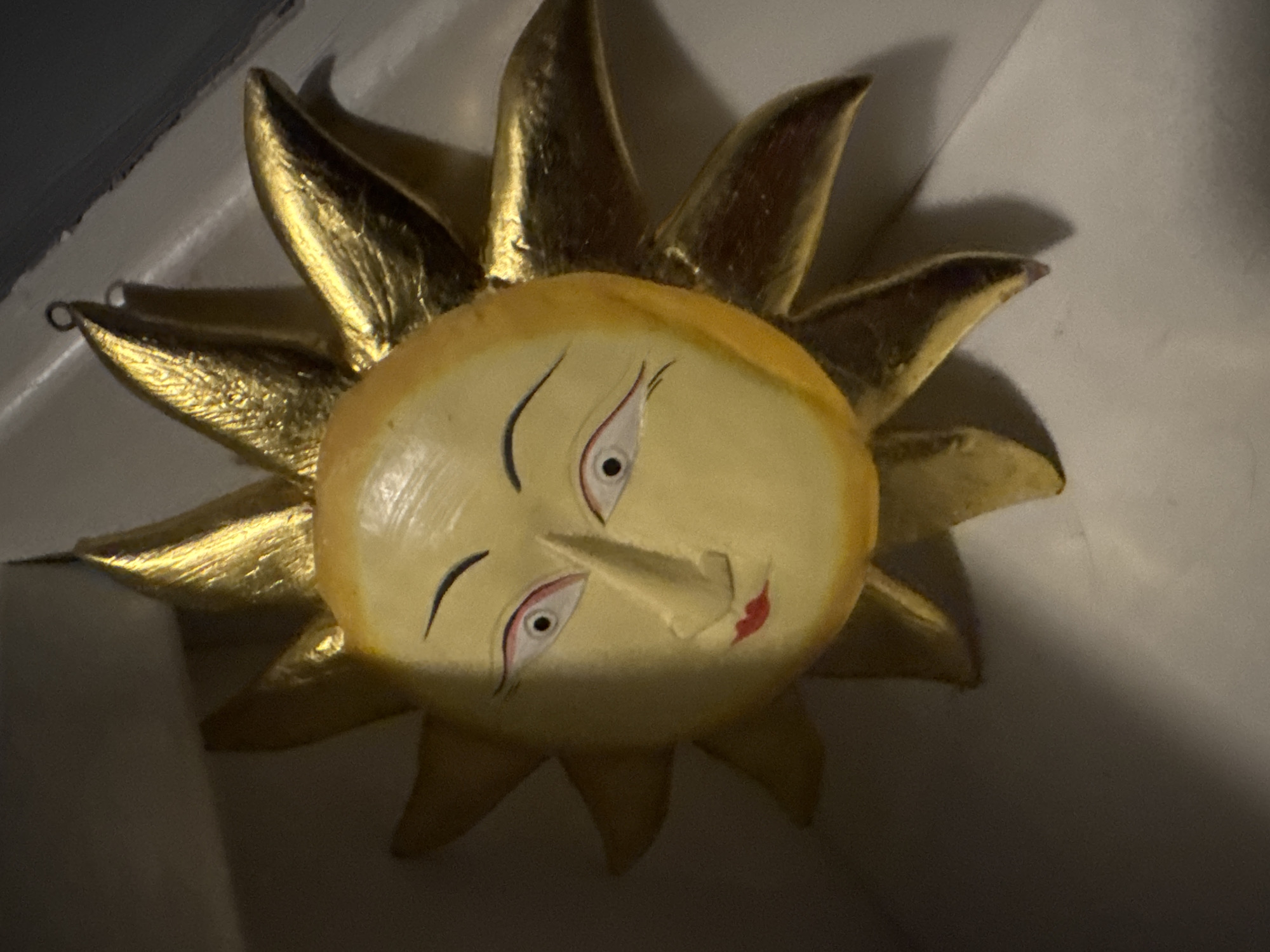 A close-up, overhead shot shows a decorative sun with a human face. The sun's face is painted yellow with red lips and black eyebrows. Its eyes are white with black pupils and red accents. The sun's rays are gold and pointed. The object is hanging from a small metal loop at the top. The background is a white surface with a dark shadow on the left.