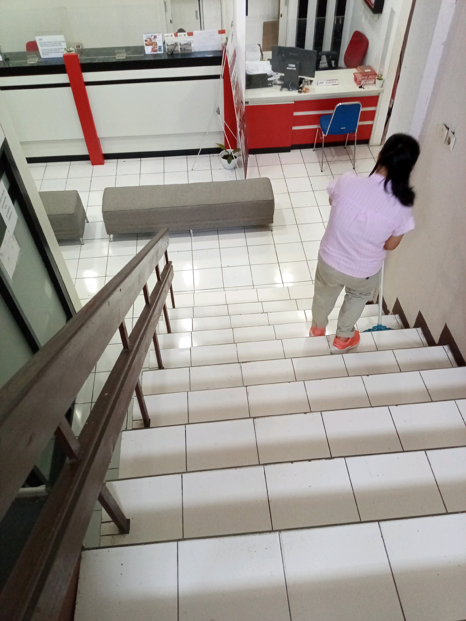 A woman with dark hair pulled back is mopping the tiled stairs of an indoor staircase. She is wearing a light pink striped shirt and light beige pants, with bright orange shoes. The staircase has a dark brown wooden railing on the left side of the frame. The stairs themselves are white ceramic tiles with dark grout lines. At the top of the stairs, visible in the background, is a reception area with a counter, a blue chair, and a computer. The scene appears to be a commercial or office building in Bitung, Indonesia.