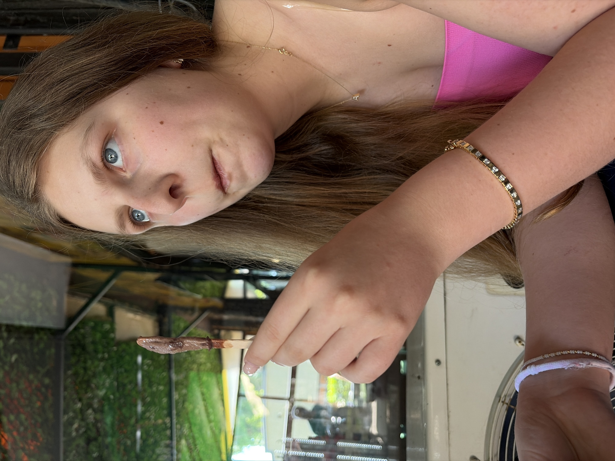 A young woman is captured in a close-up, rotated portrait, looking directly at the viewer with an inquisitive or surprised expression. She has long, light brown hair, fair skin with a few freckles, and piercing blue eyes. Her lips are slightly parted.
She is wearing a vibrant pink top or dress, visible on her upper body. Around her neck, she wears a delicate gold chain necklace with a small, minimalist pendant. On her left wrist, a gold-colored bracelet adorned with alternating dark and light rectangular stones is visible. Her right wrist features two additional bracelets: one appearing to be a white, fabric-like band and another made of small beads.
In her right hand, she holds a thin wooden stick with a dollop of dark, semi-solid food, likely a dessert like chocolate or Nutella, on its end. She seems to be holding it up, perhaps to offer or to eat.
The background is softly out of focus but suggests an outdoor or semi-outdoor setting, possibly a cafe or restaurant in Burgas, Bulgaria. Green foliage, resembling a hedge or planted wall, is visible, along with reflections from windows or glass panels. The overall lighting indicates it is daytime. There is no discernible text within the image.