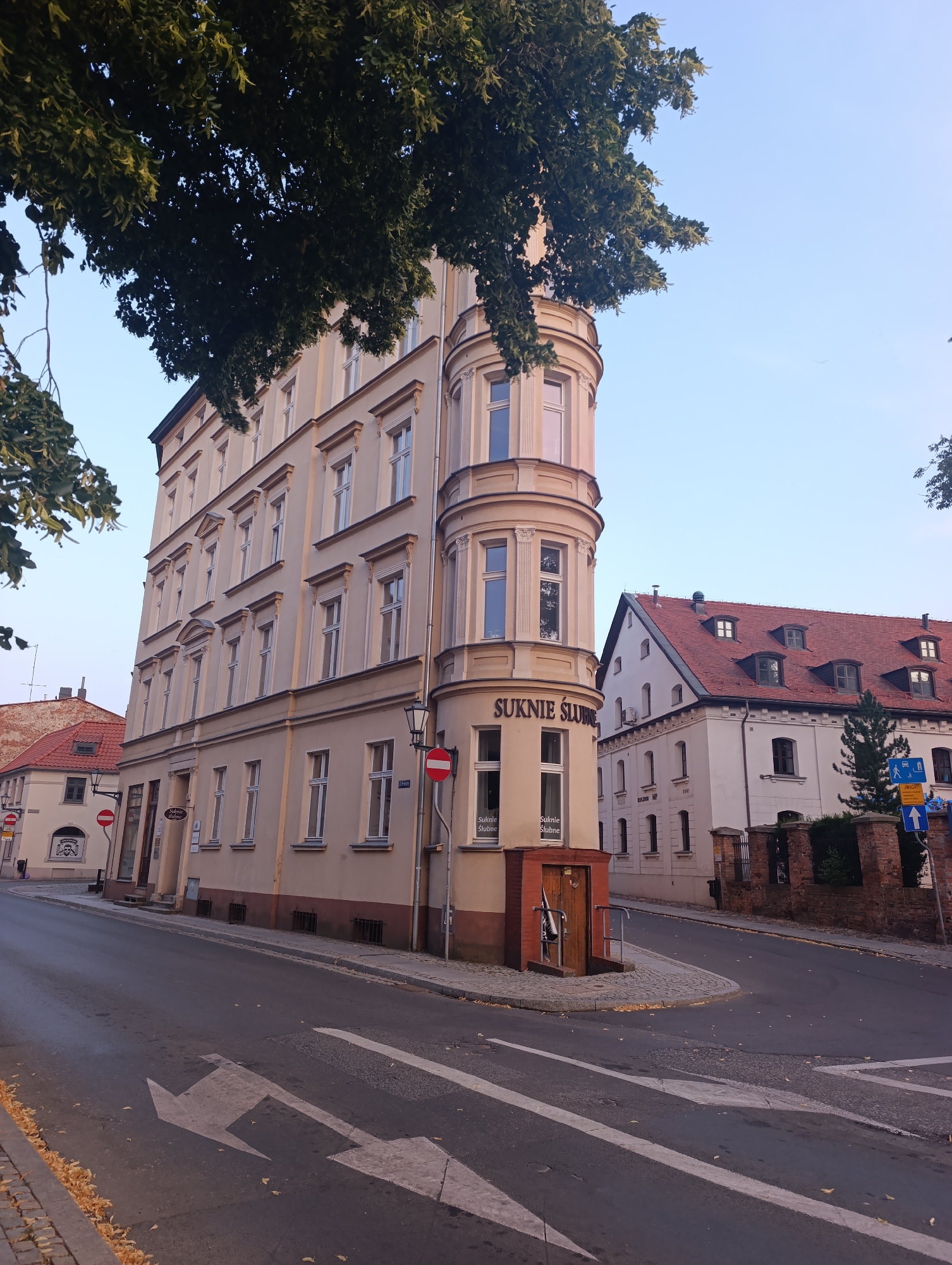 In the city of Toruń, Poland, a street view shows a cream-colored, multi-story building with a distinct rounded corner featuring a bay window. The building has numerous rectangular windows with white frames. Above the bay window, signage reads "SUKNIE ŚLUBNE." To the right, a white building with a red-tiled roof and dormer windows is visible. A street with traffic markings, including arrows and a pedestrian crossing, dominates the foreground. Fallen leaves are scattered on the roadside and sidewalk. The sky is clear and blue.