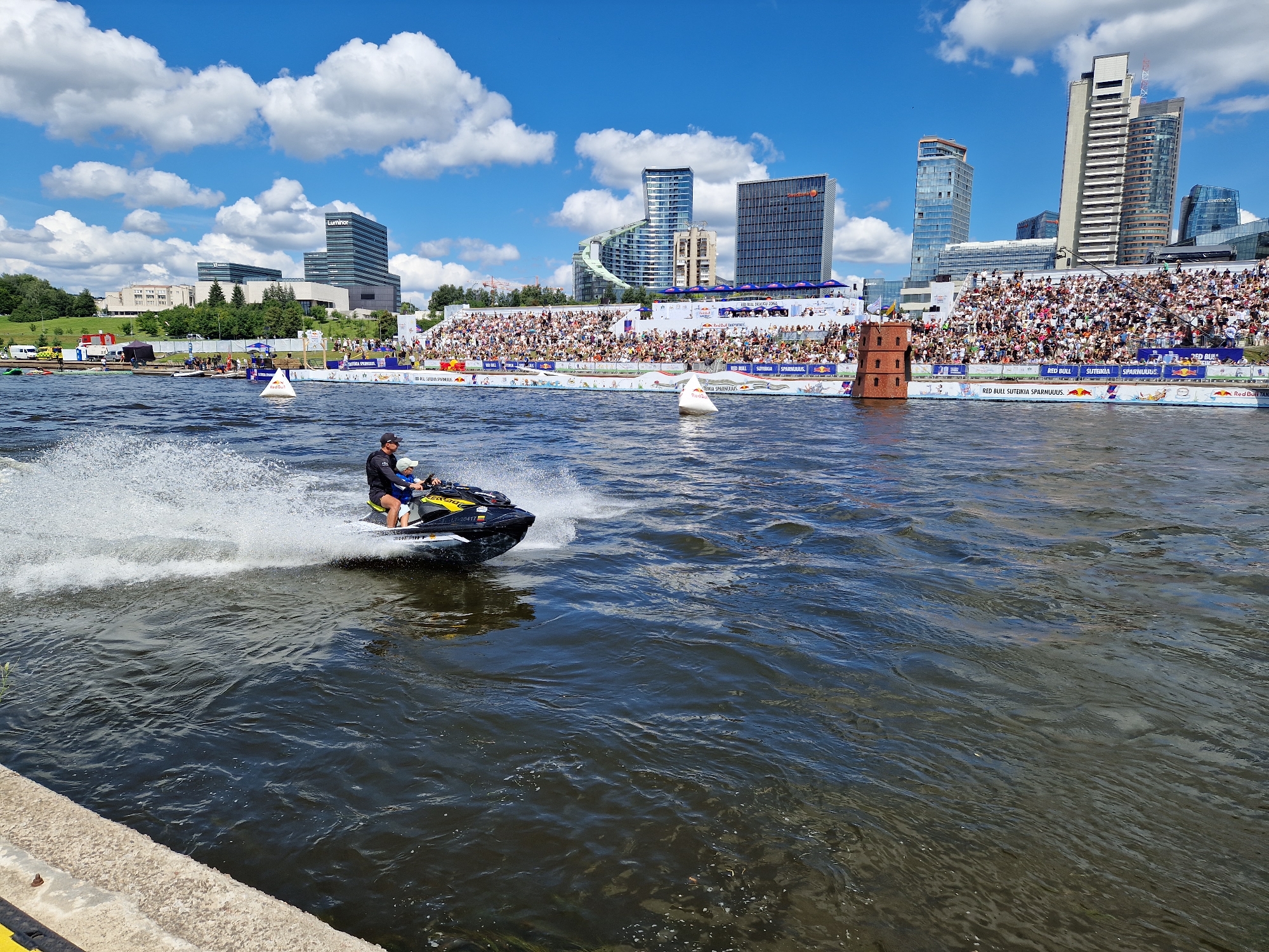 This is a wide-angle, eye-level shot of a sunny day event on the Neris River in Vilnius, Lithuania. In the foreground, a jet ski carrying two individuals is moving from left to right, creating a spray of water. The jet ski is black and yellow and has "SEA-DOO" written on it. The rider is wearing a dark shirt and hat, and the passenger appears to be a child in a blue shirt and life jacket.
In the mid-ground, the riverbank is lined with a tiered seating area filled with spectators. A white inflatable buoy with a Red Bull logo is floating in the water near the seating. Further back, a small, multi-level brick tower stands in the water, possibly a navigational marker or a decorative element. Beyond the spectators, the Vilnius cityscape is visible with modern skyscrapers and office buildings, including the distinctive curved "Europa Tower" and the "Luminor" building. The sky is blue with fluffy white clouds. The overall scene suggests an outdoor sporting event or festival taking place on the river.
