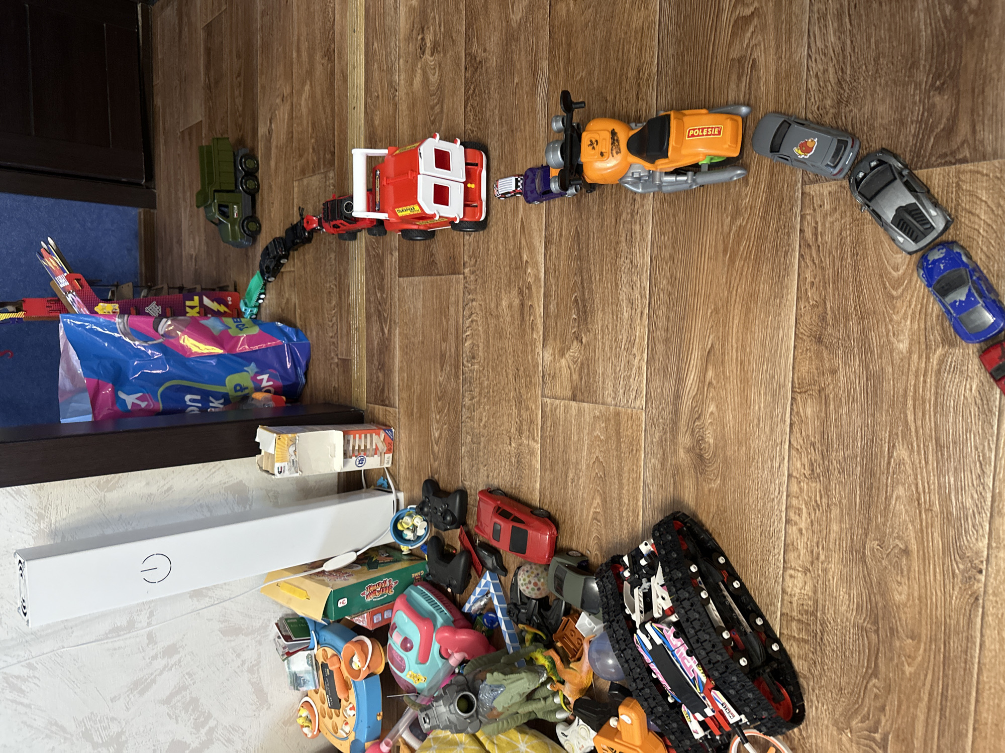 The image depicts a collection of children's toys scattered on a wooden floor. In the foreground, a toy tank with black treads and colorful decals is visible. To the right, a series of toy vehicles, including cars and a motorcycle, are arranged in a line, forming a winding path across the floor. Among these are a red truck, a green toy truck, and several smaller cars in various colors. In the background, a tall white object with a power symbol is standing upright. Other scattered toys, including a blue plastic bucket and a green dinosaur figurine, are also present. The scene appears to be an indoor play area, likely a child's room. There are no people or discernible actions in the image, only a static arrangement of toys. The location is not specified in the image.